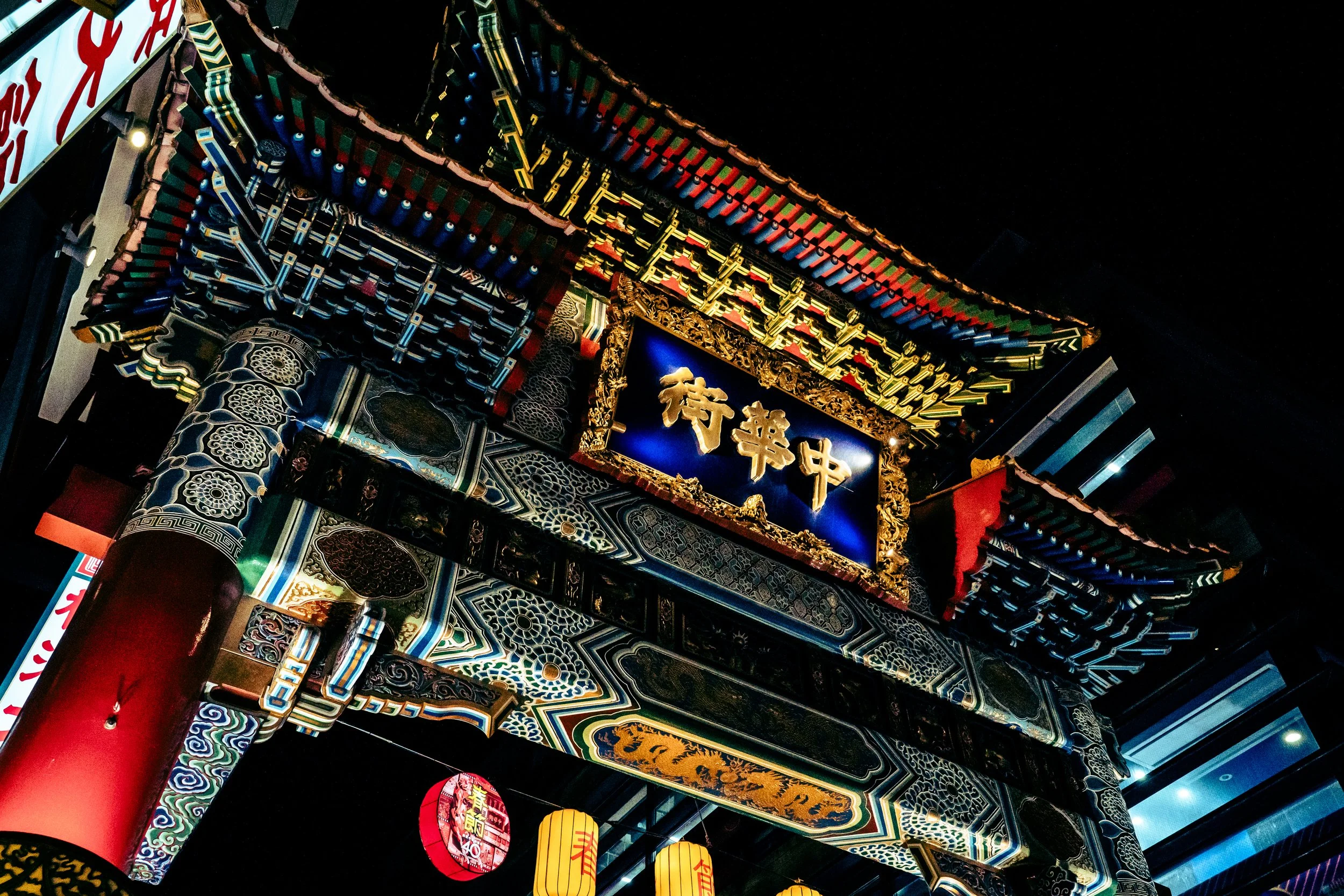 Nighttime view of a traditional Chinese or Japanese decorated gate with ornate designs, bright colors, illuminated signs, and hanging lanterns.