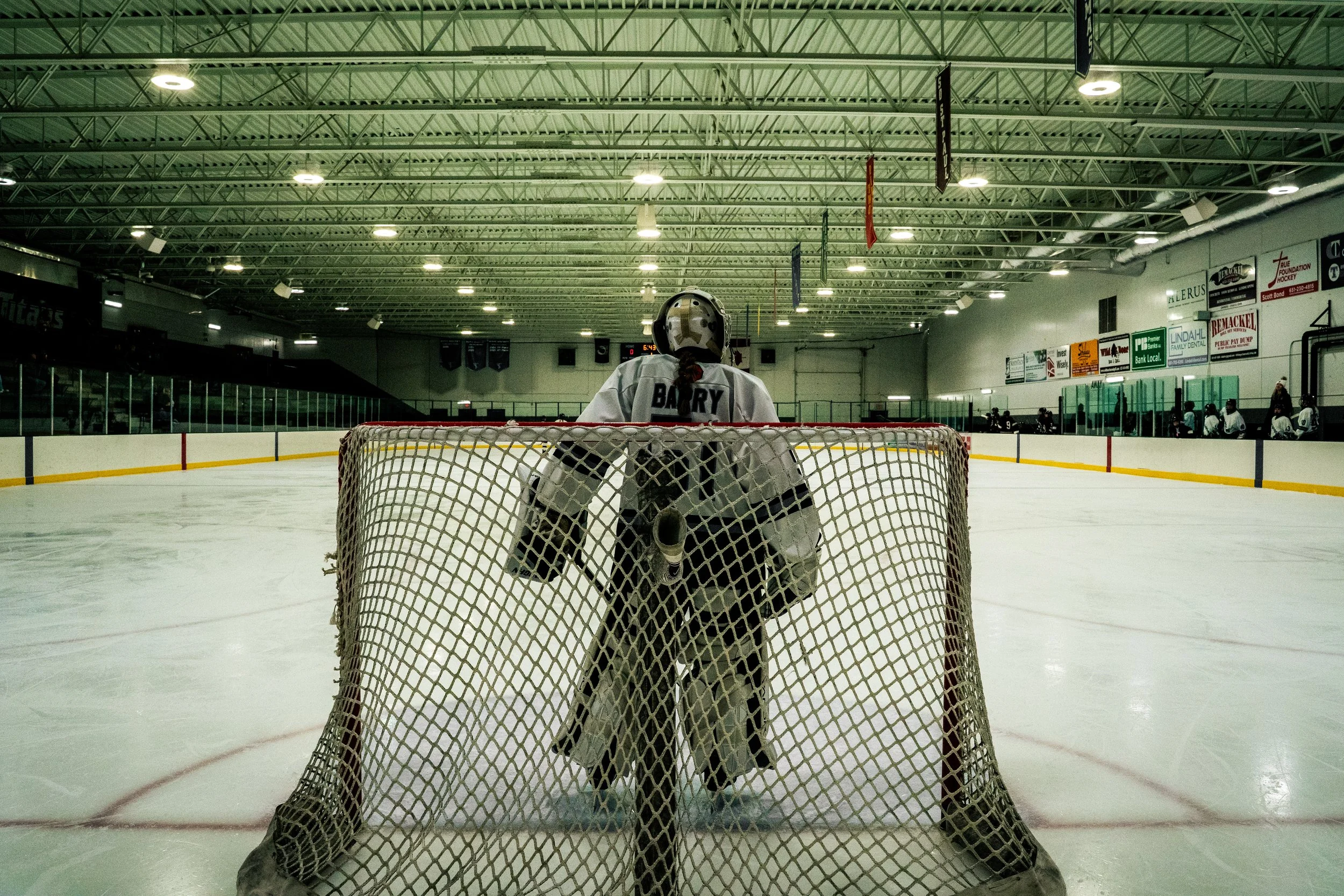 Ice hockey goalie standing in front of the net on an ice rink, facing away, wearing a white jersey with the name 'BARRY' and protective gear, with players and advertisements visible in the background.