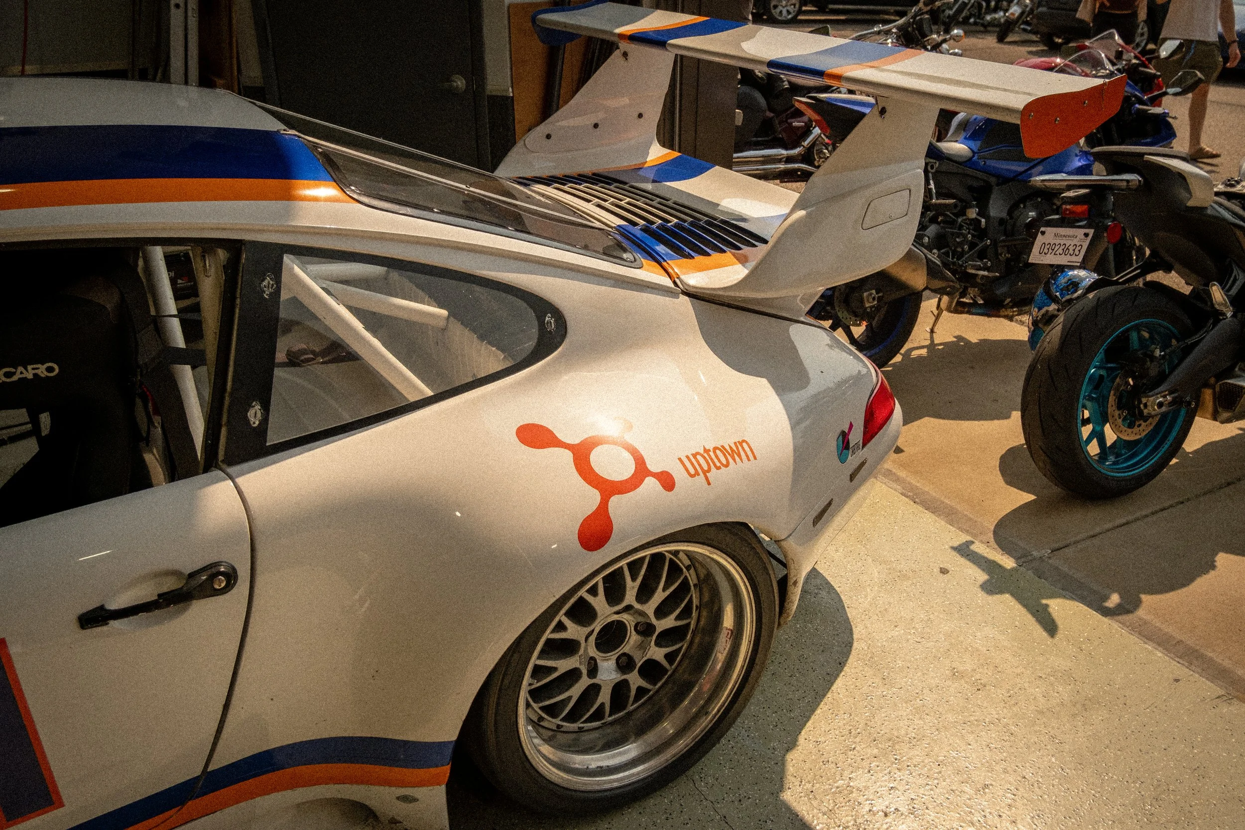 Close-up image of a white race car with blue and orange accents, showing the front and part of the side, parked indoors with a wheel visible.