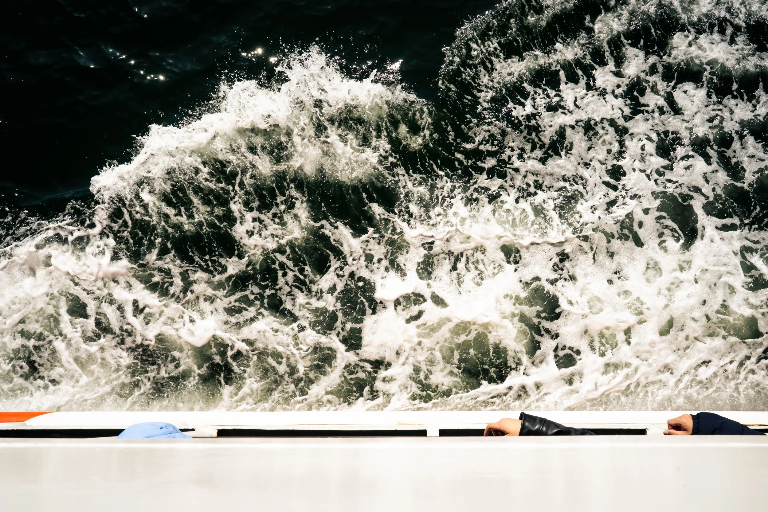 view from a boat showing dark ocean water with white foam and waves, with the boat's edge and two hands visible at the bottom of the image