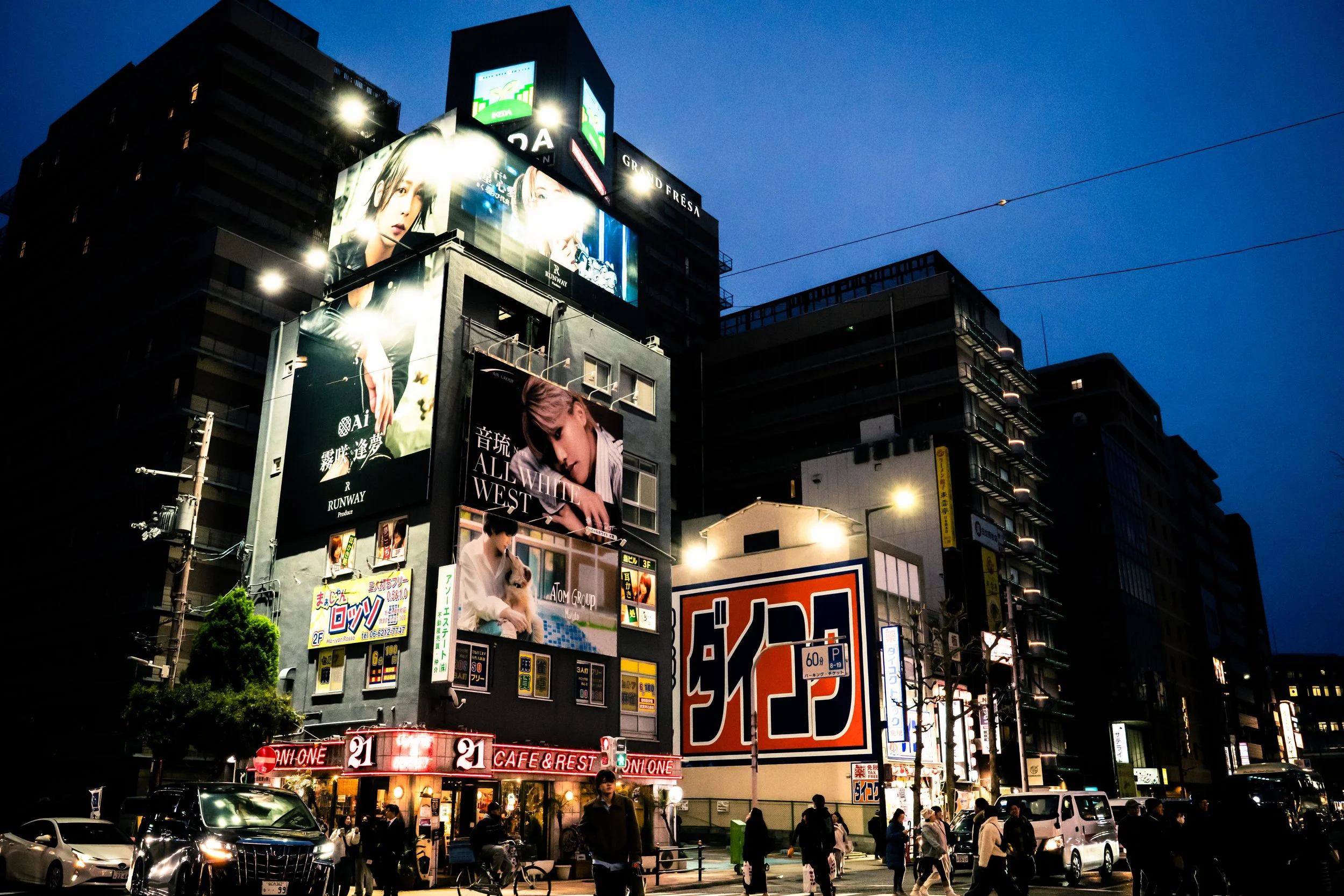 Nighttime city scene with large illuminated billboards on tall buildings, displaying various advertisements, and people walking on the street below.