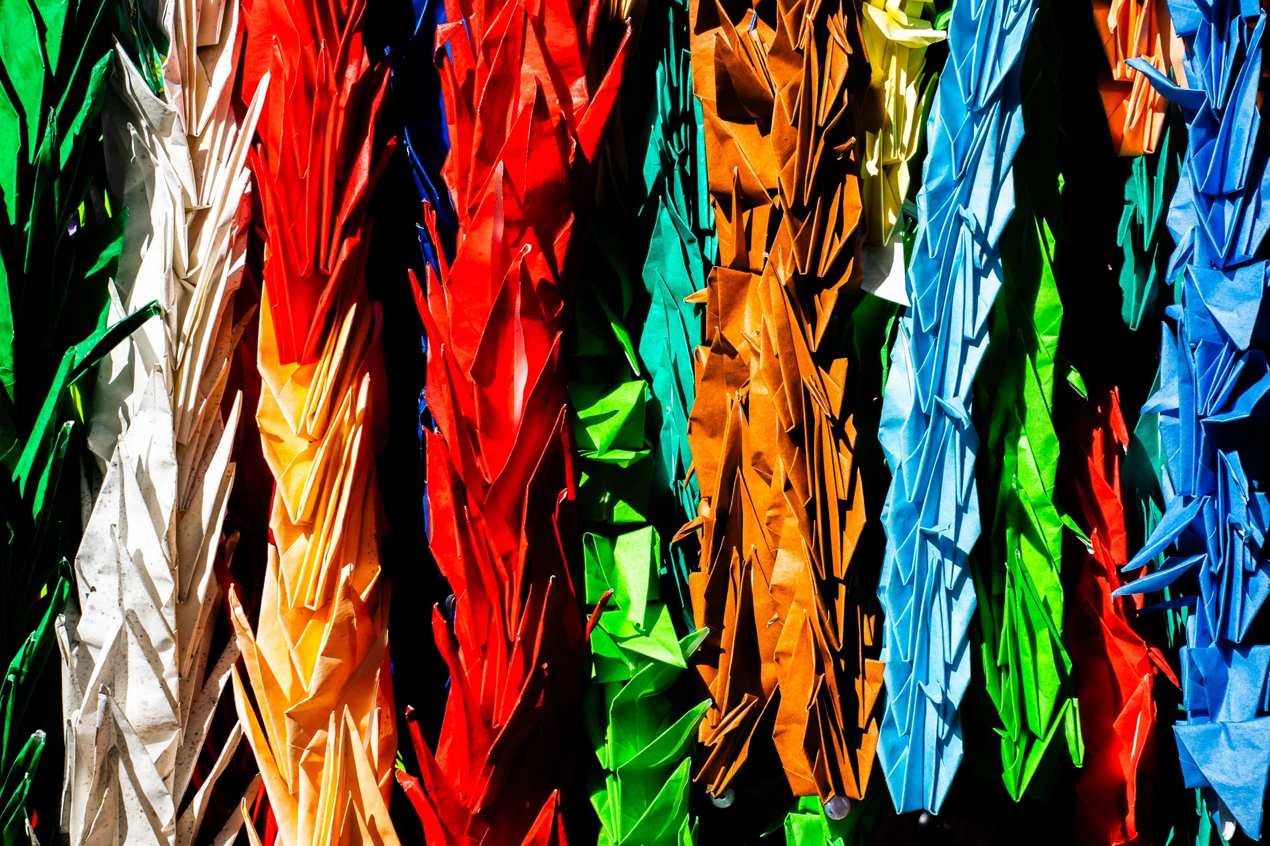 Close-up of numerous colorful origami paper cranes hanging in clusters against a dark background.