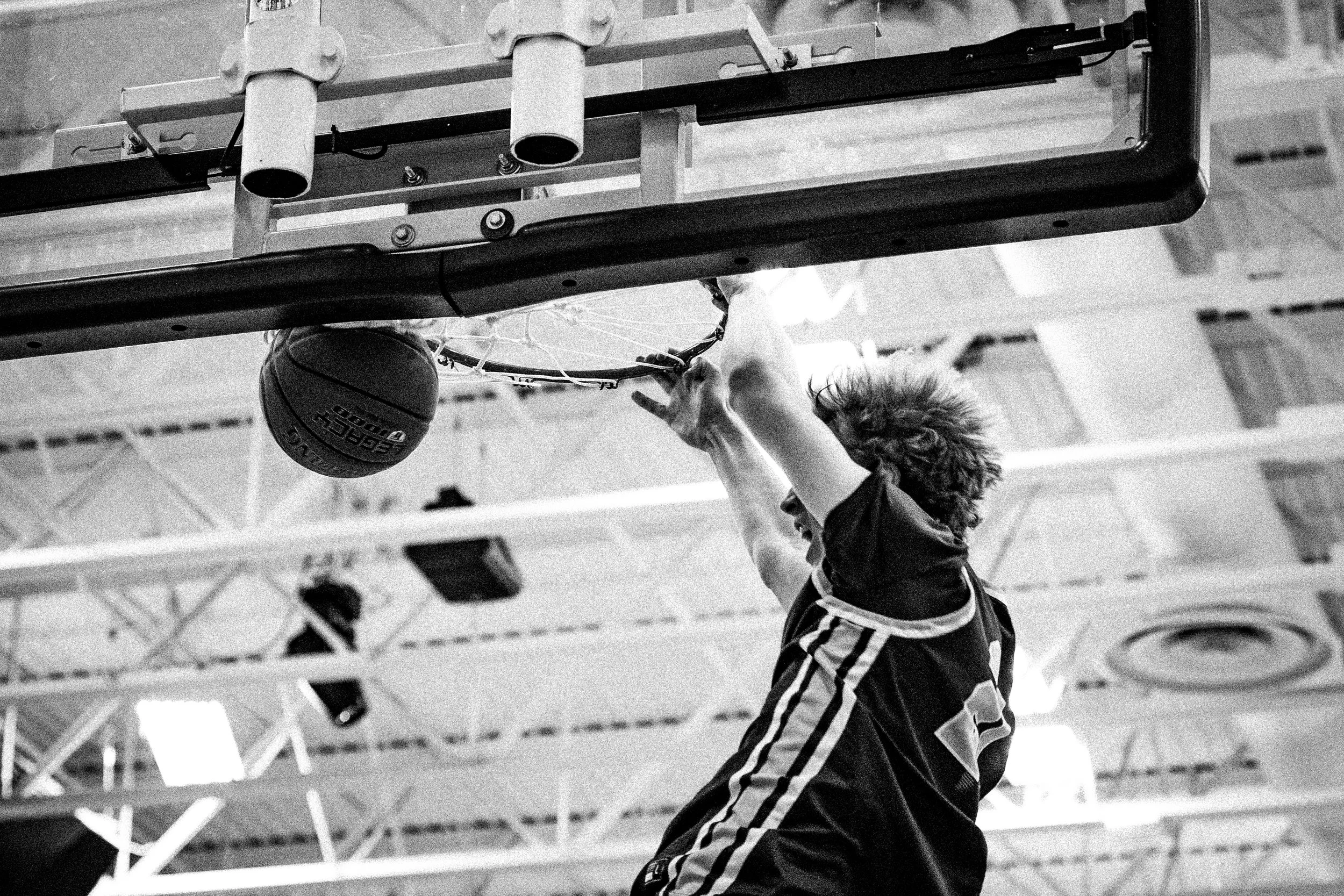A young man wearing a striped sports jacket and gloves practicing basketball in a gymnasium, reaching up towards the hoop to make a shot. The gym has a high ceiling with lighting fixtures.