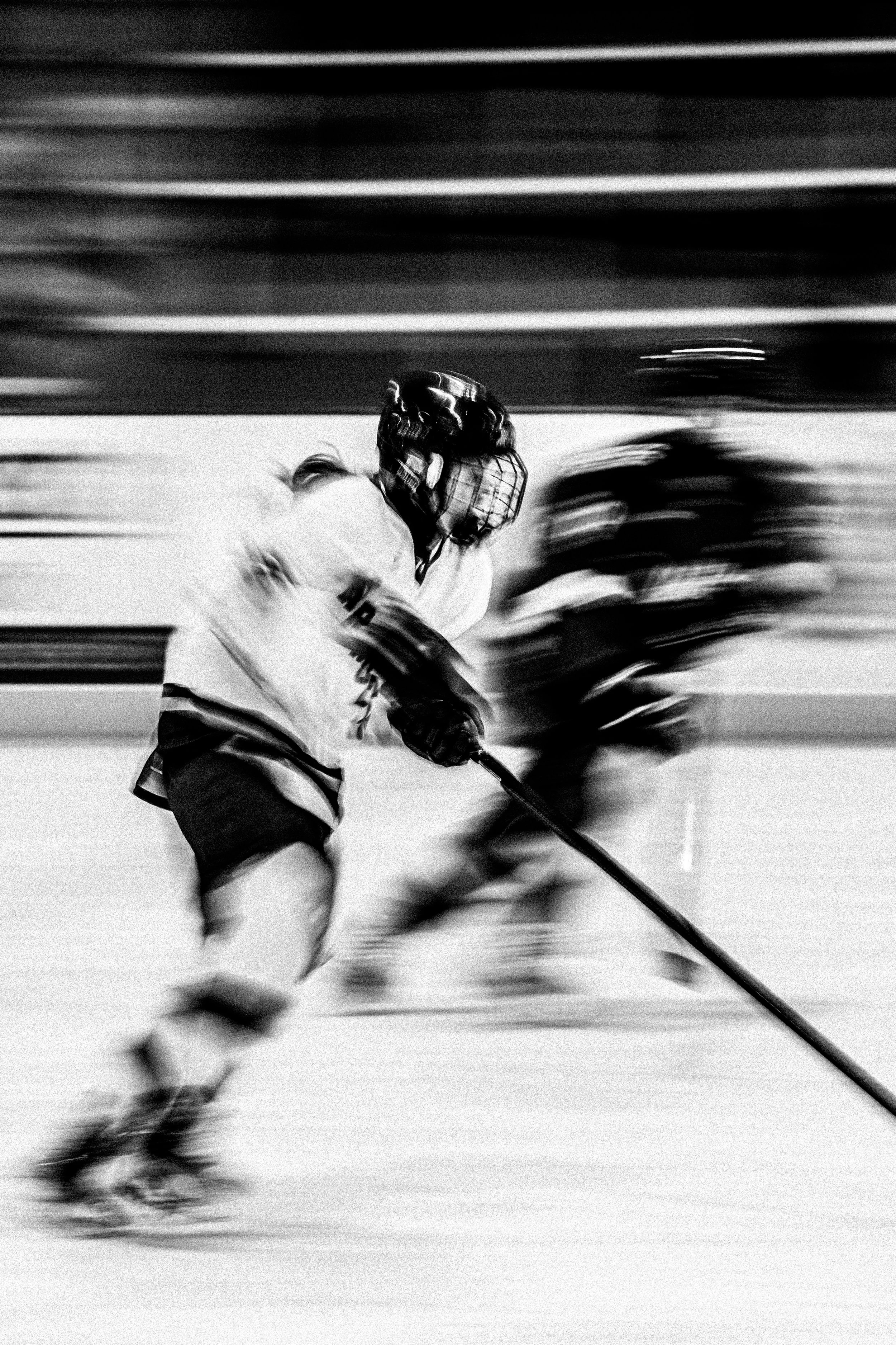 Black and white photo of ice hockey players in motion on the rink, with one player in focus wearing a helmet and sports gear.