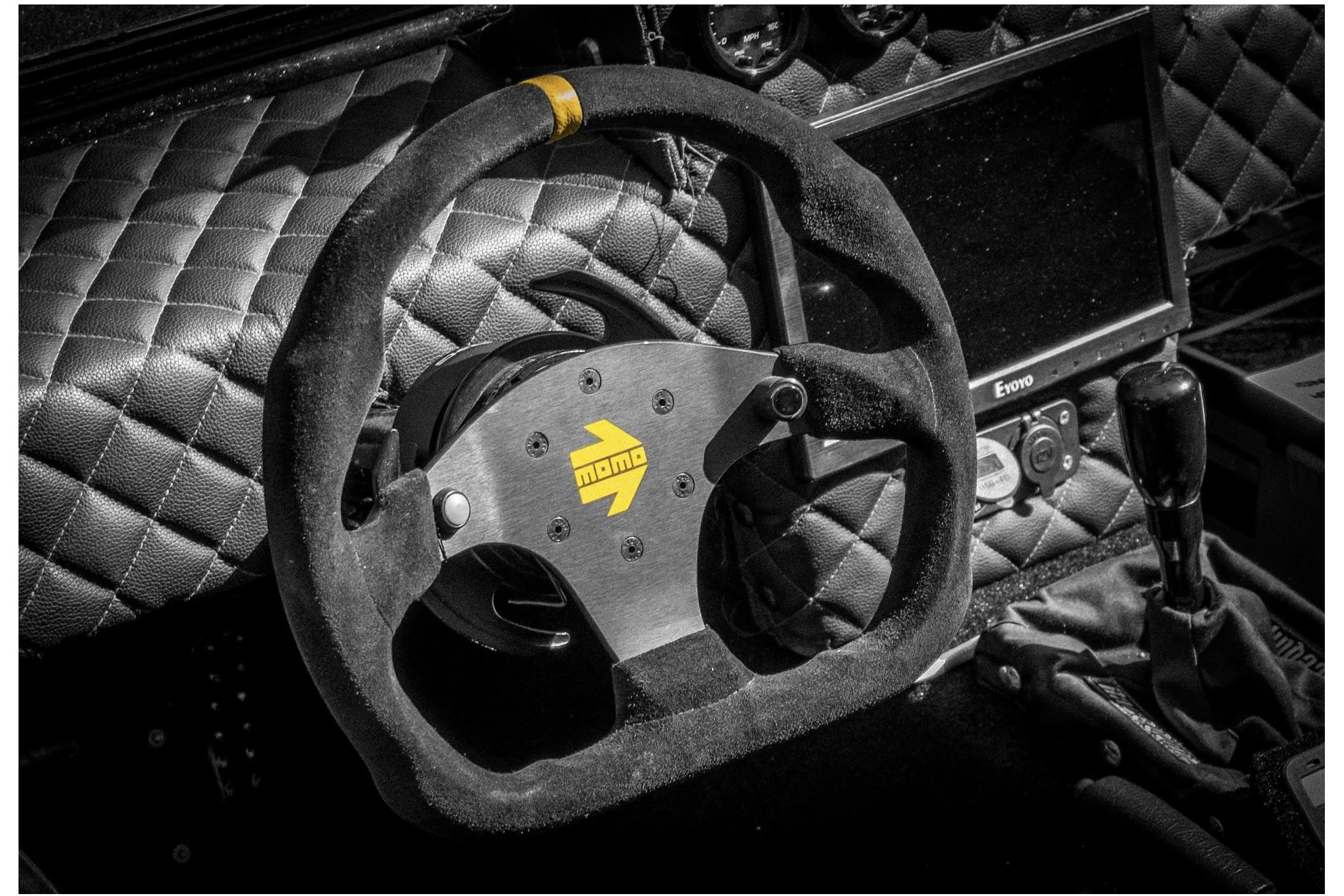 A racing car simulator interior featuring a quilted black leather seat, a black steering wheel with a yellow logo, and various gauges and controls.