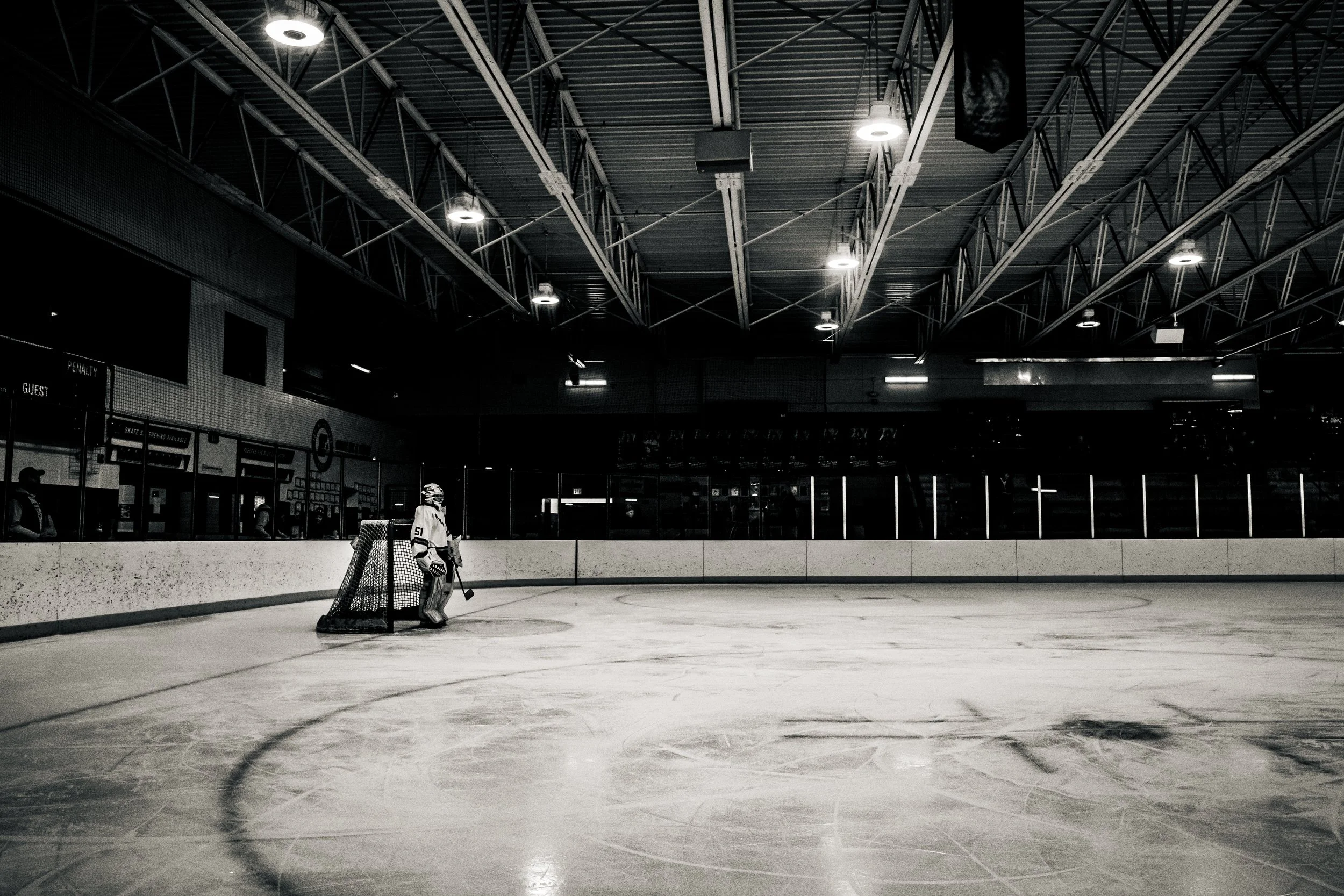 An empty indoor ice hockey rink with a single goalie standing near the goal. The ceiling has industrial lighting and metal beams, with no spectators visible.