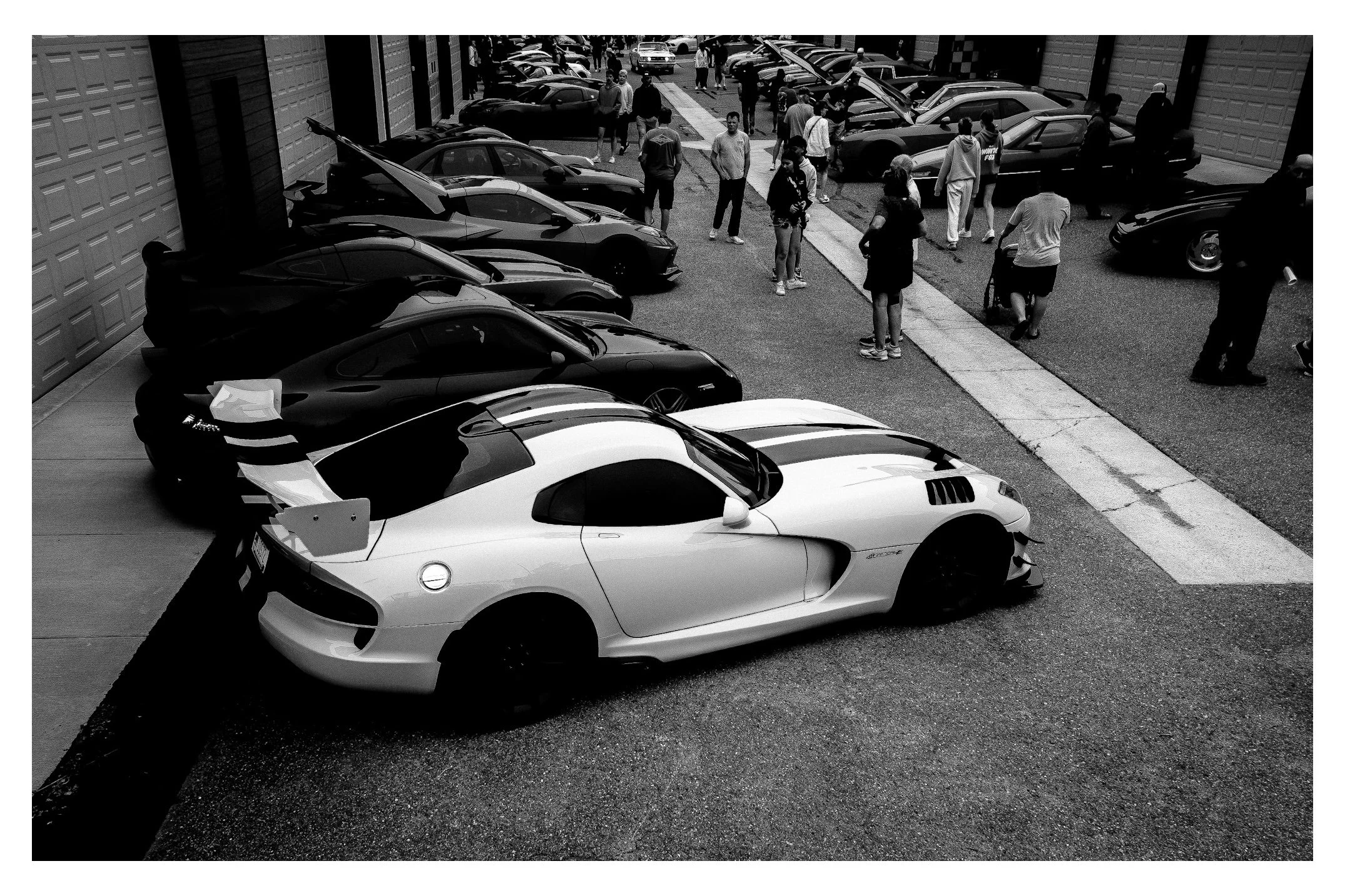 Exterior view of a parking lot filled with luxury sports cars, with a group of people walking and mingling in the background.