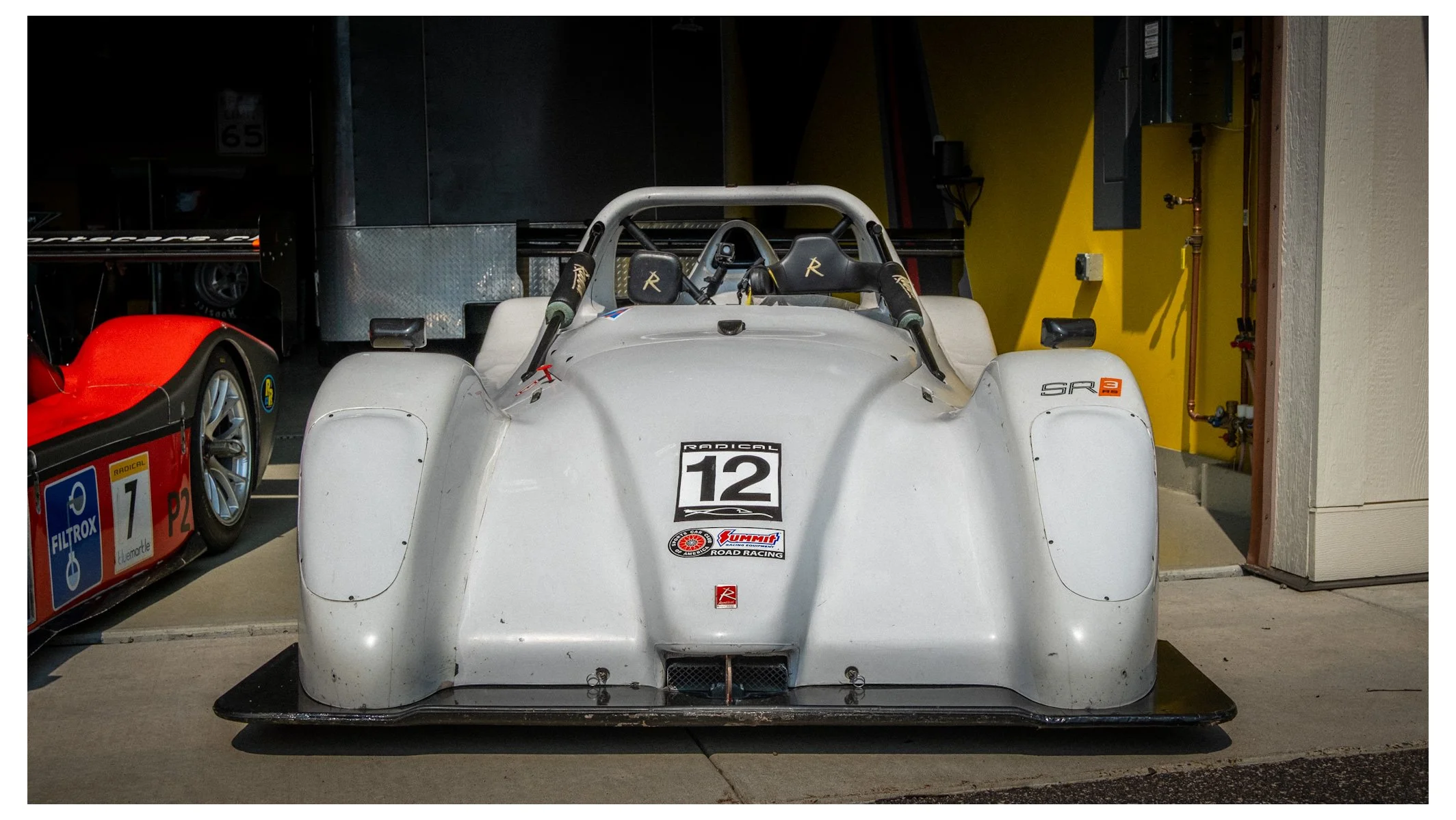 A white race car, number 12, parked indoors with a yellow wall and various pipes on the right side.