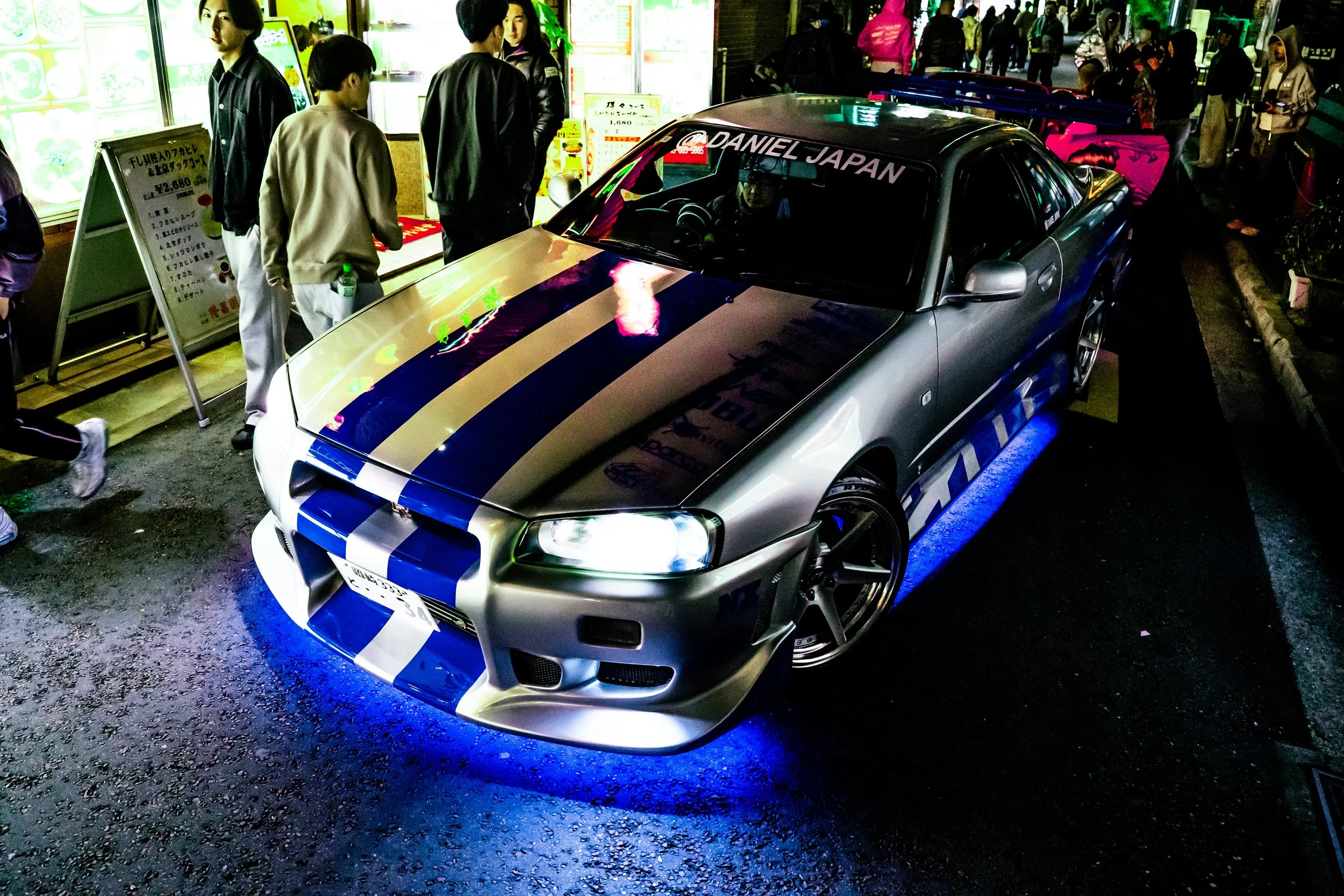 A silver and blue rally-style car with illuminated blue underglow, parked on a street at night, surrounded by people walking and interacting, some standing near the car and others in the background.