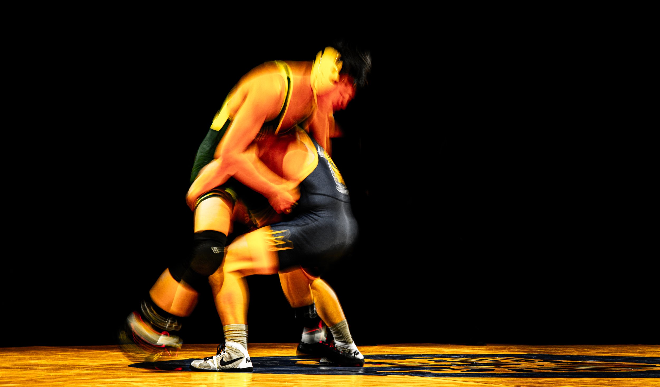 Two wrestlers in action on a wrestling mat, with one executing a move on the other, against a dark background.