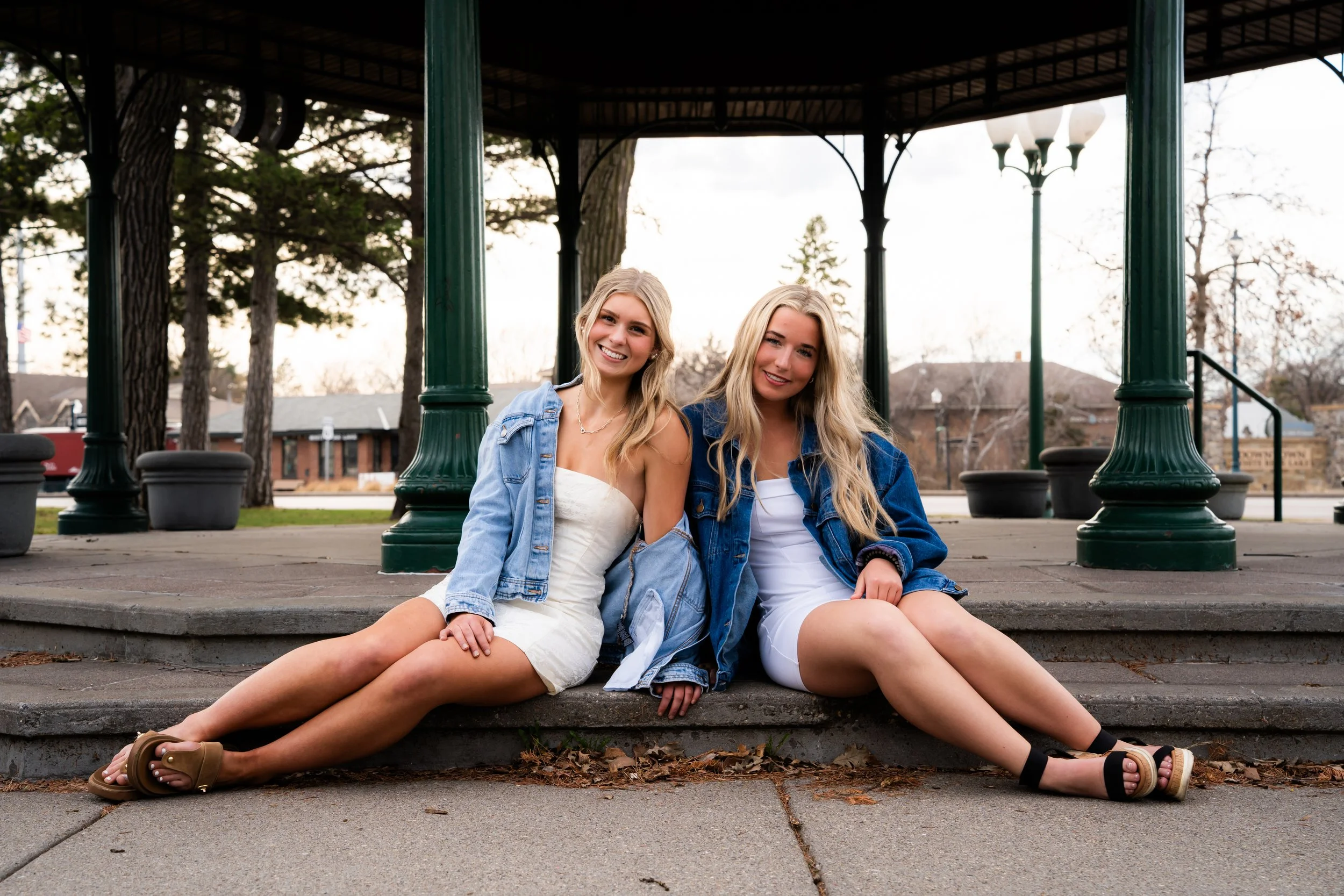 Two young women sitting on steps in a park under a pavilion, smiling at the camera. One is wearing a white dress and tan sandals, the other a denim jacket and white dress, both with long blonde hair and casual summer outfits.