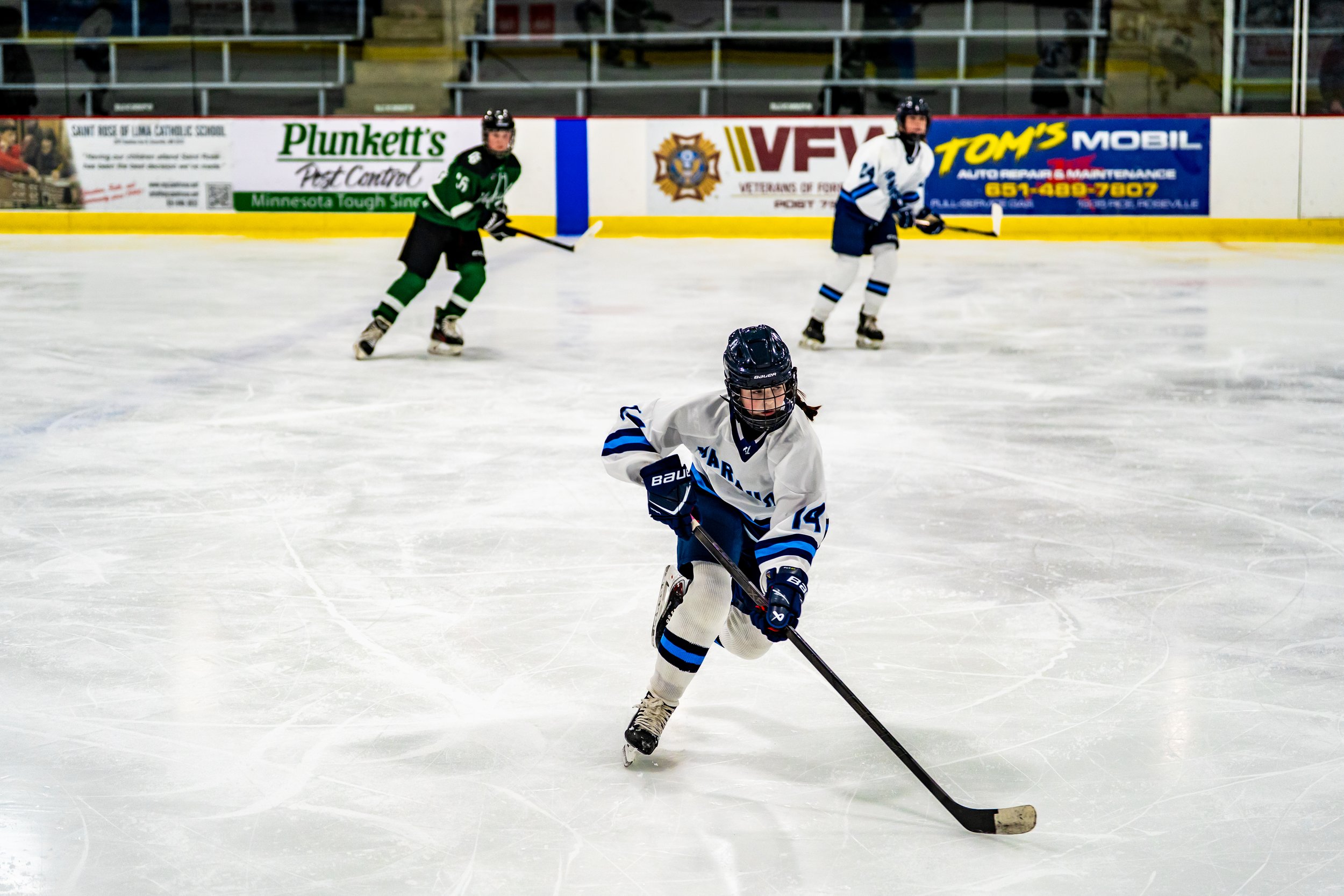 Ice hockey players in action on the rink, with three players visible wearing different team jerseys, and advertisements along the boards in the background.