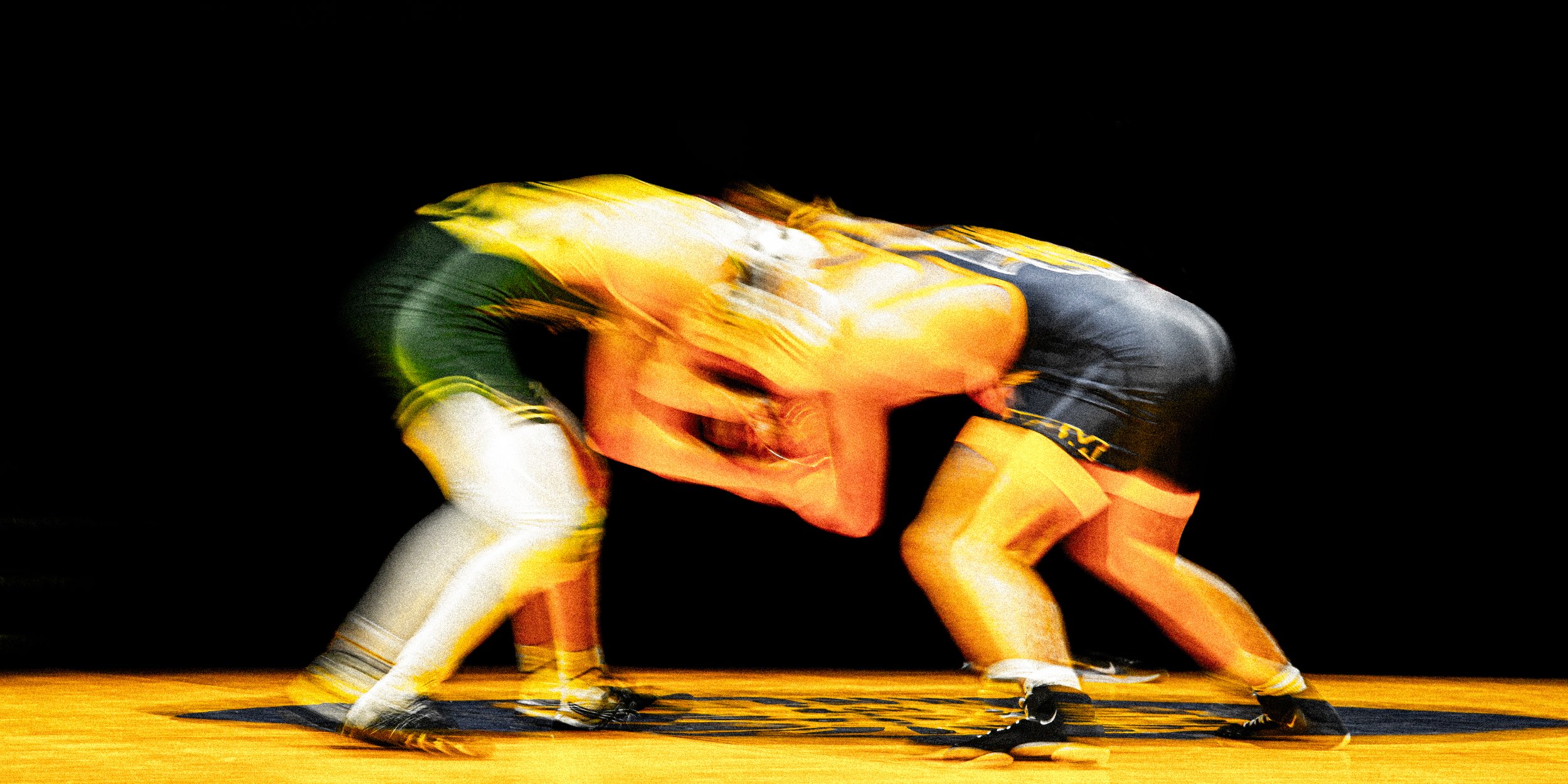 Two wrestlers engaged in a match on a wrestling mat, with one wrestler in black and yellow gear and the other in yellow and white gear, captured mid-motion.