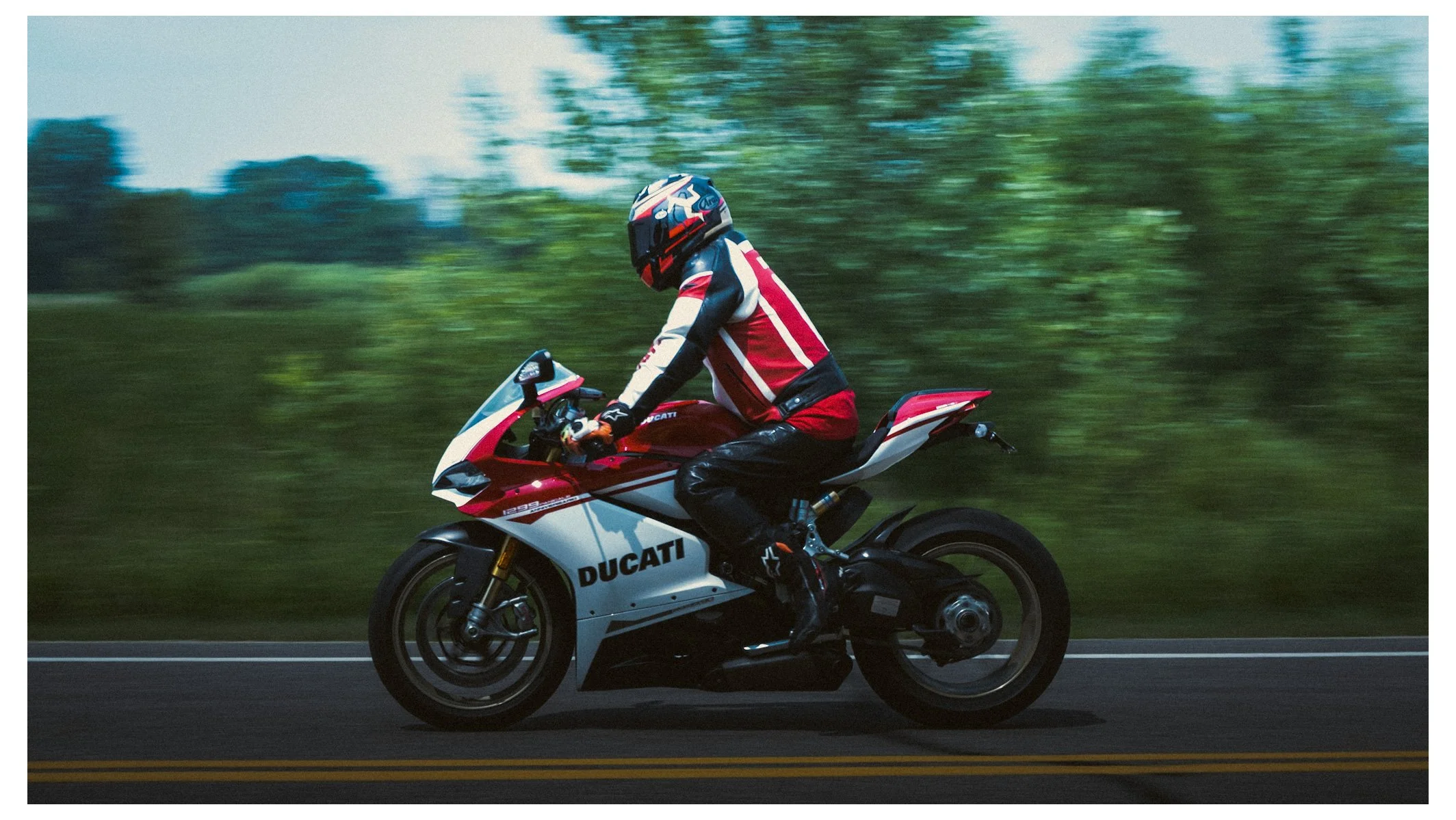 A person riding a Ducati sport motorcycle on a road with greenery in the background, wearing a helmet and a racing suit.