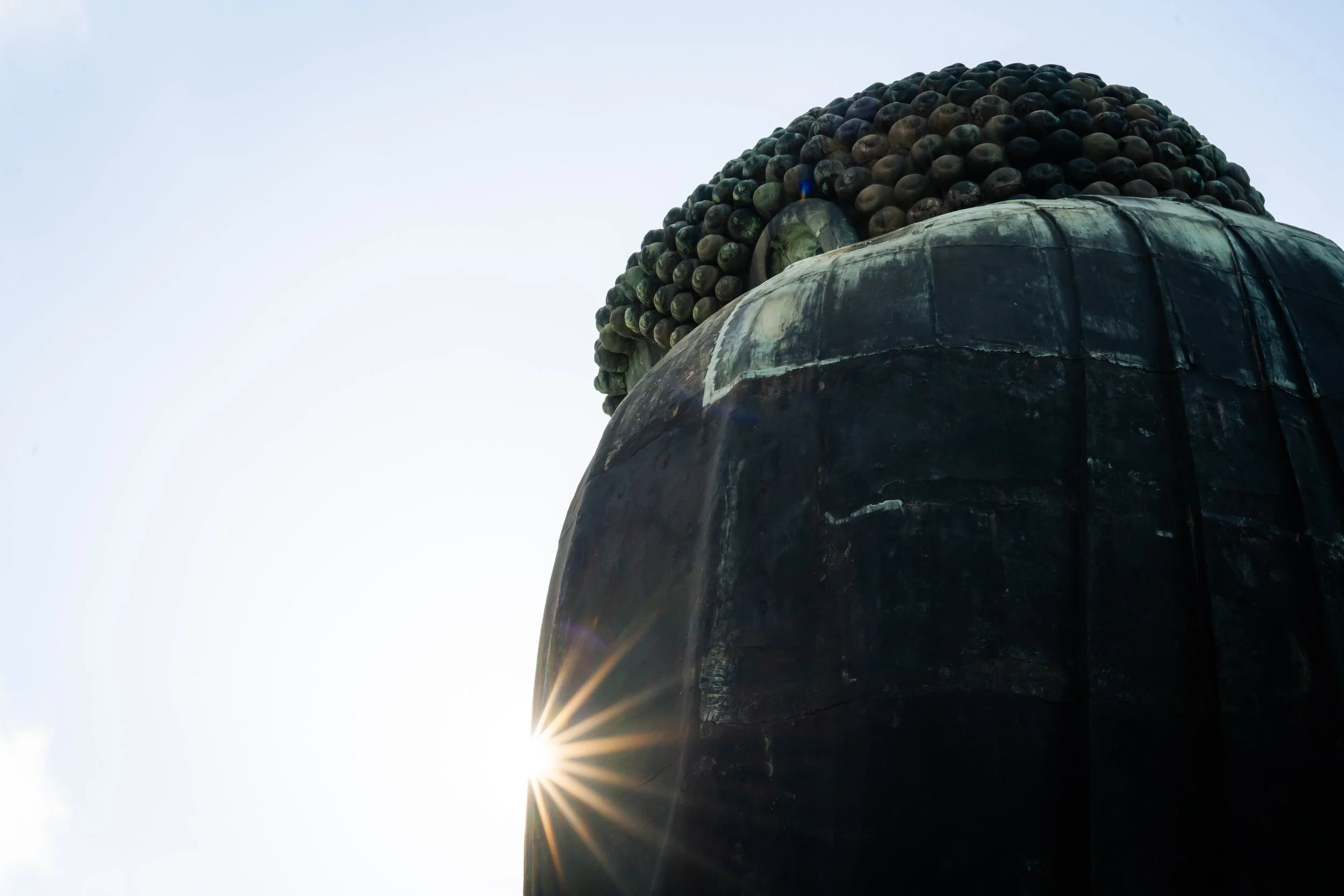 A close-up side view of a large statue's head with a serene expression, focusing on the face and hair, with the sun shining behind the statue creating a starburst effect and the sky in the background.