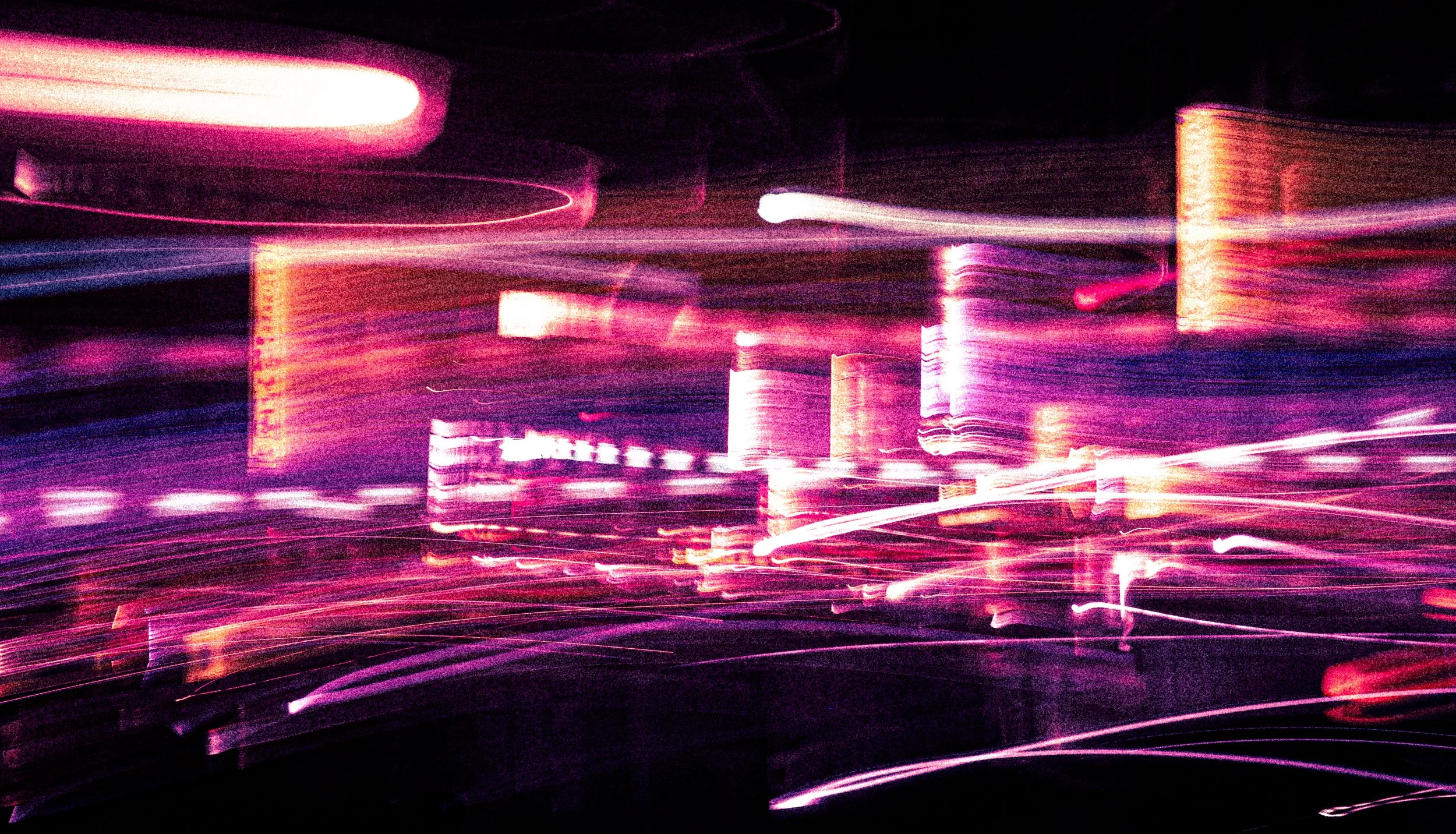 Blurred cityscape at night with streaks of pink, purple, and white lights and tall buildings.