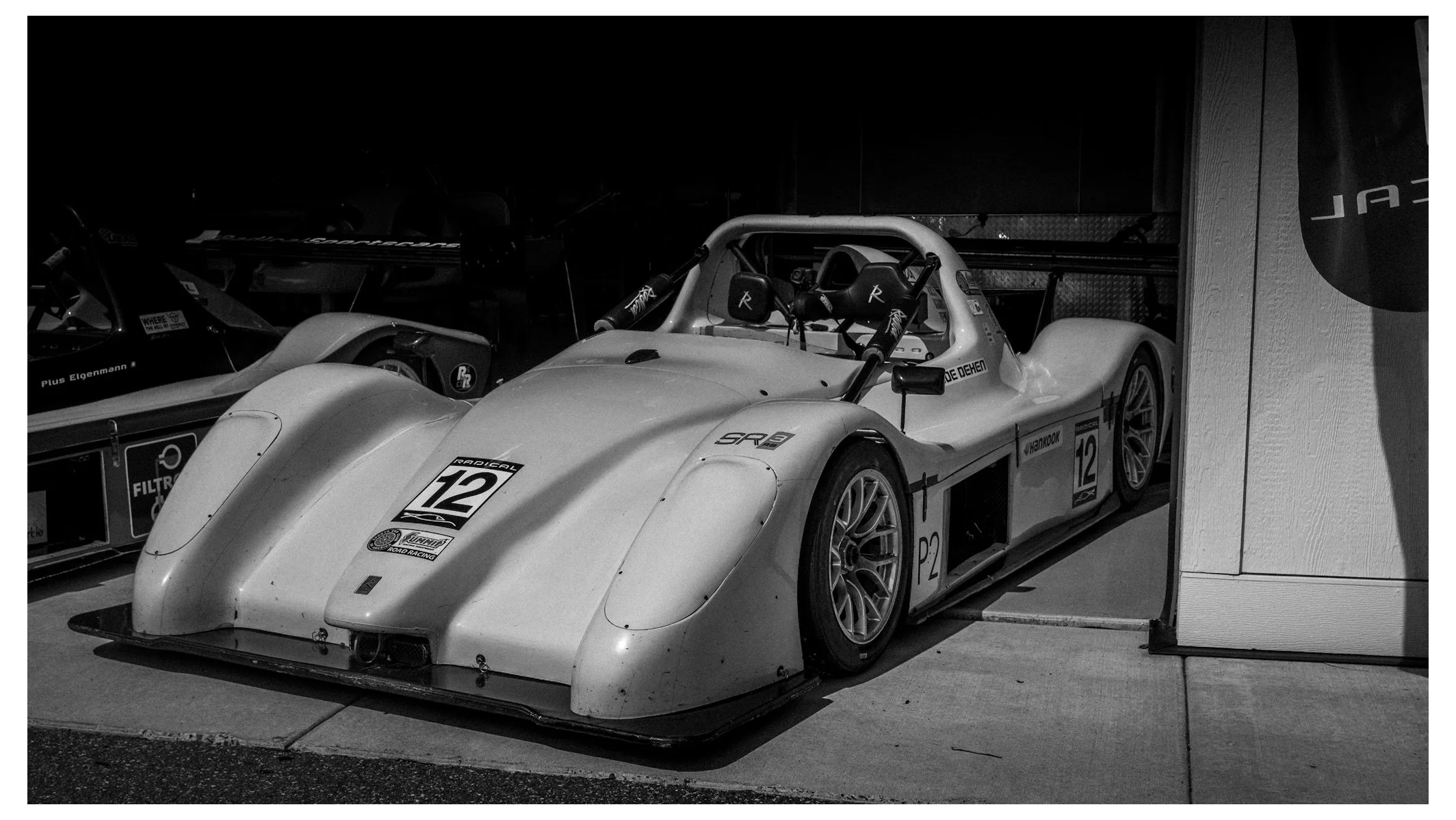 Black and white photo of a race car with the number 12 on its front, parked indoors next to another race car partially visible to its left.