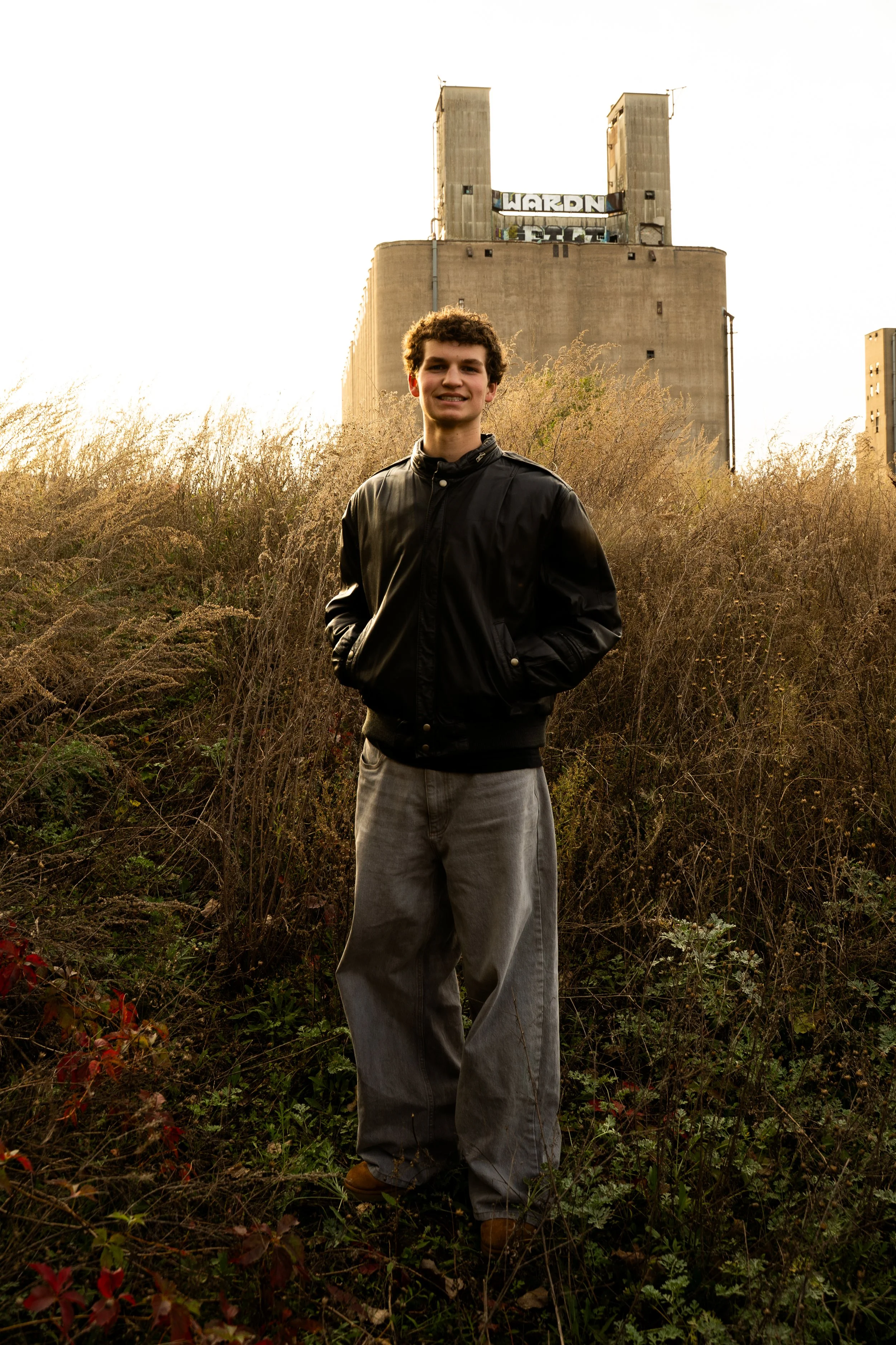 Young man with curly hair wearing a black jacket and gray pants standing outdoors among tall dry grass and plants, with a large concrete building in the background and bright sky above.