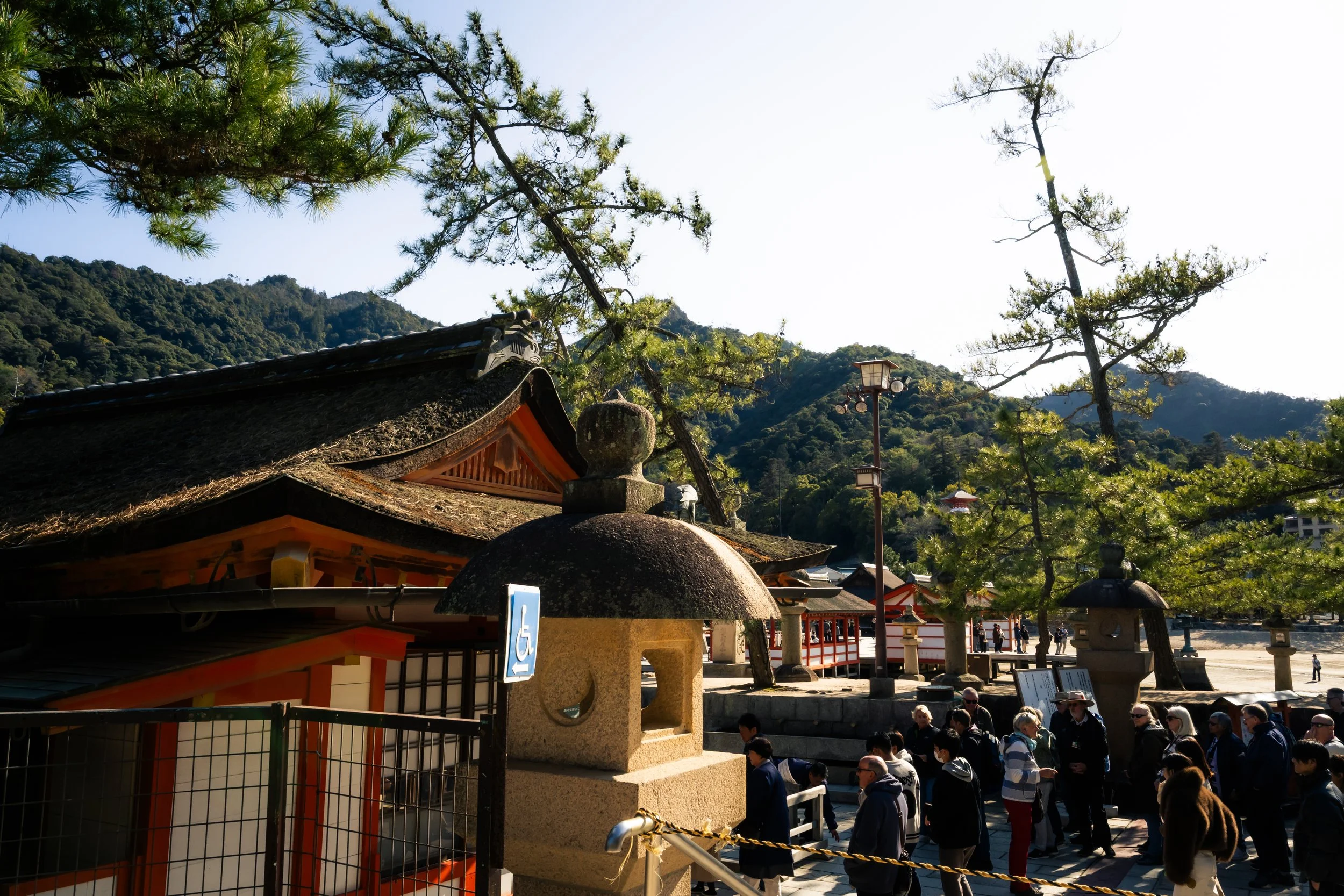 A group of people standing in line at a shrine or temple, with traditional Japanese architecture, trees, and mountains in the background.
