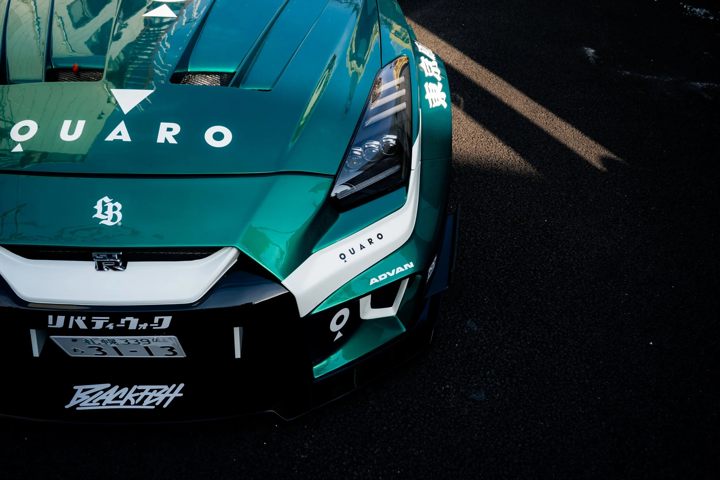 Close-up of a teal sports car with decal stickers, Japanese license plate, and decals reading QUARQ, ADVAN, and BLACKRAY, parked on a dark pavement.