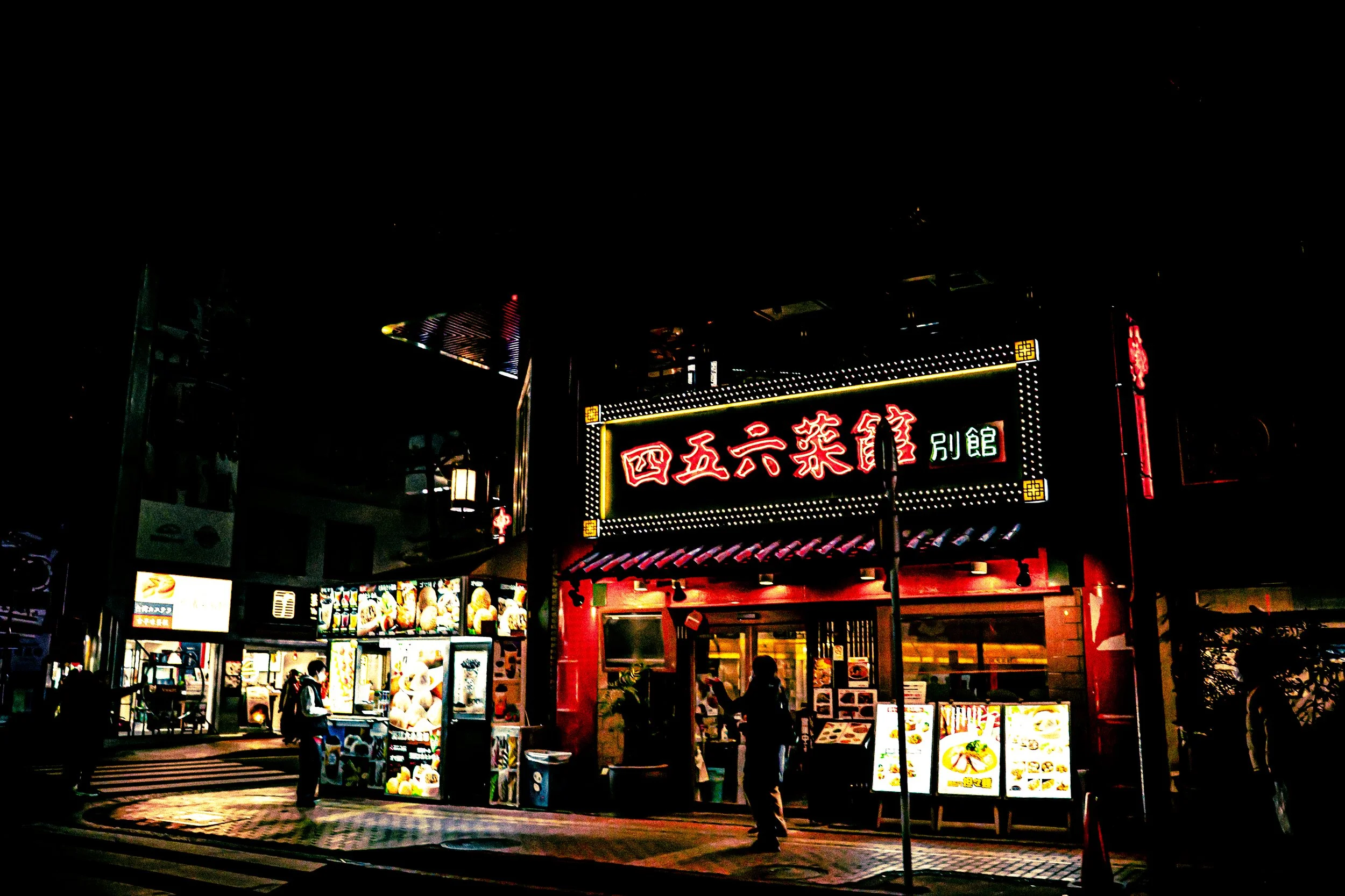 Night scene of a brightly lit Asian restaurant with neon signs and menu displays, with people walking on the sidewalk.