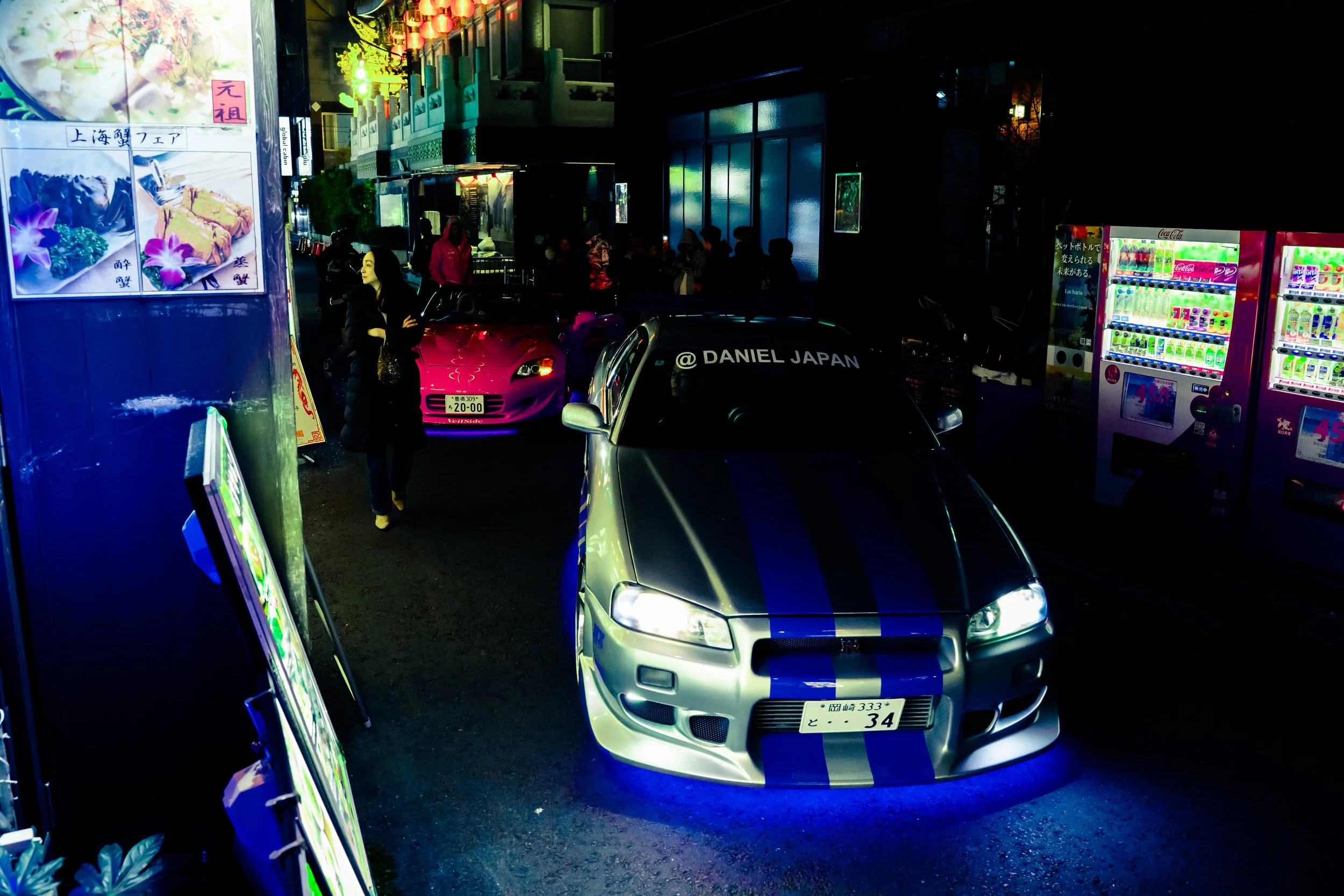 Night scene of a busy street with parked cars, vending machines, illuminated signs, and people walking near a building with traditional Japanese architecture.