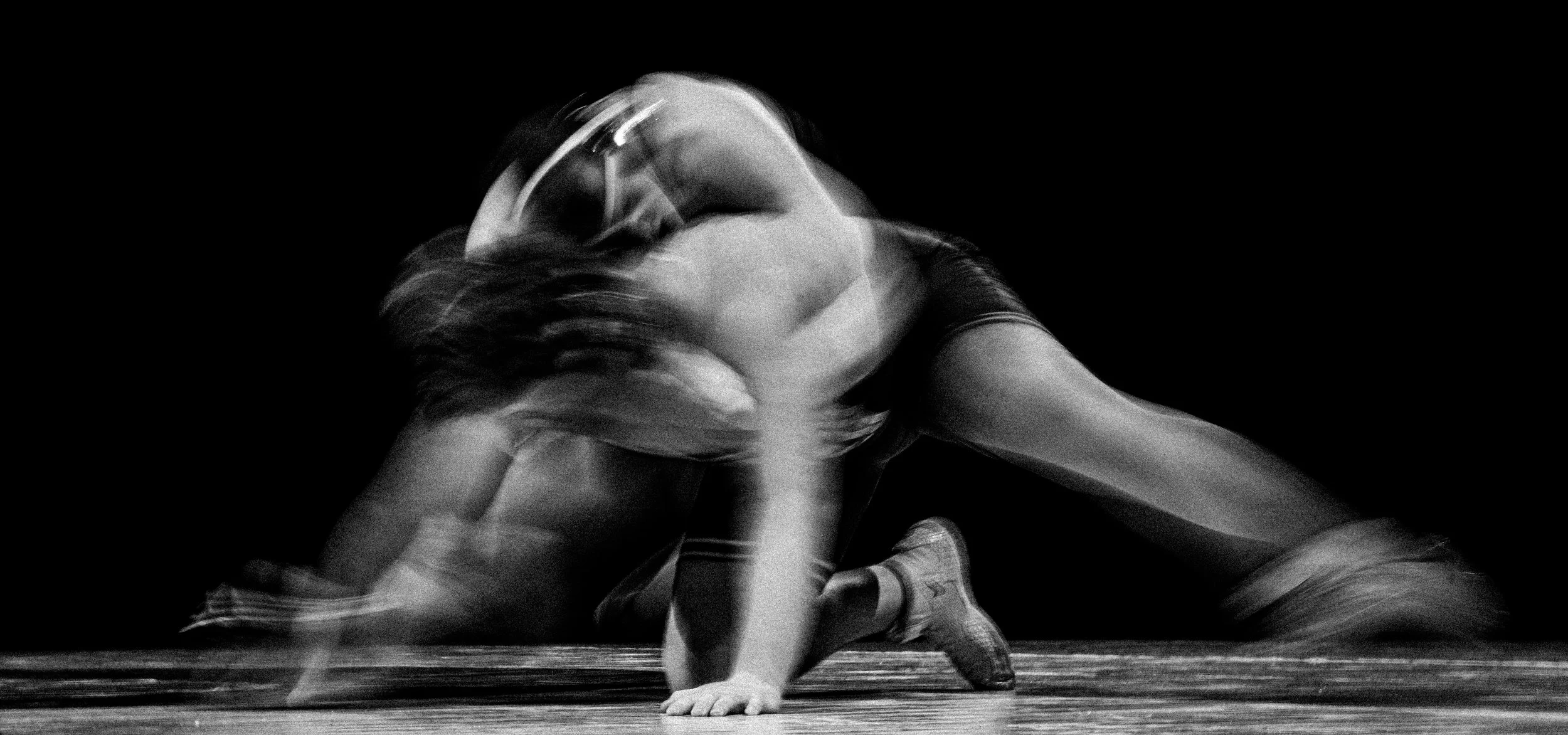 A black and white photo of a person in a dynamic pose, possibly dancing or performing, on a stage with a dark background.