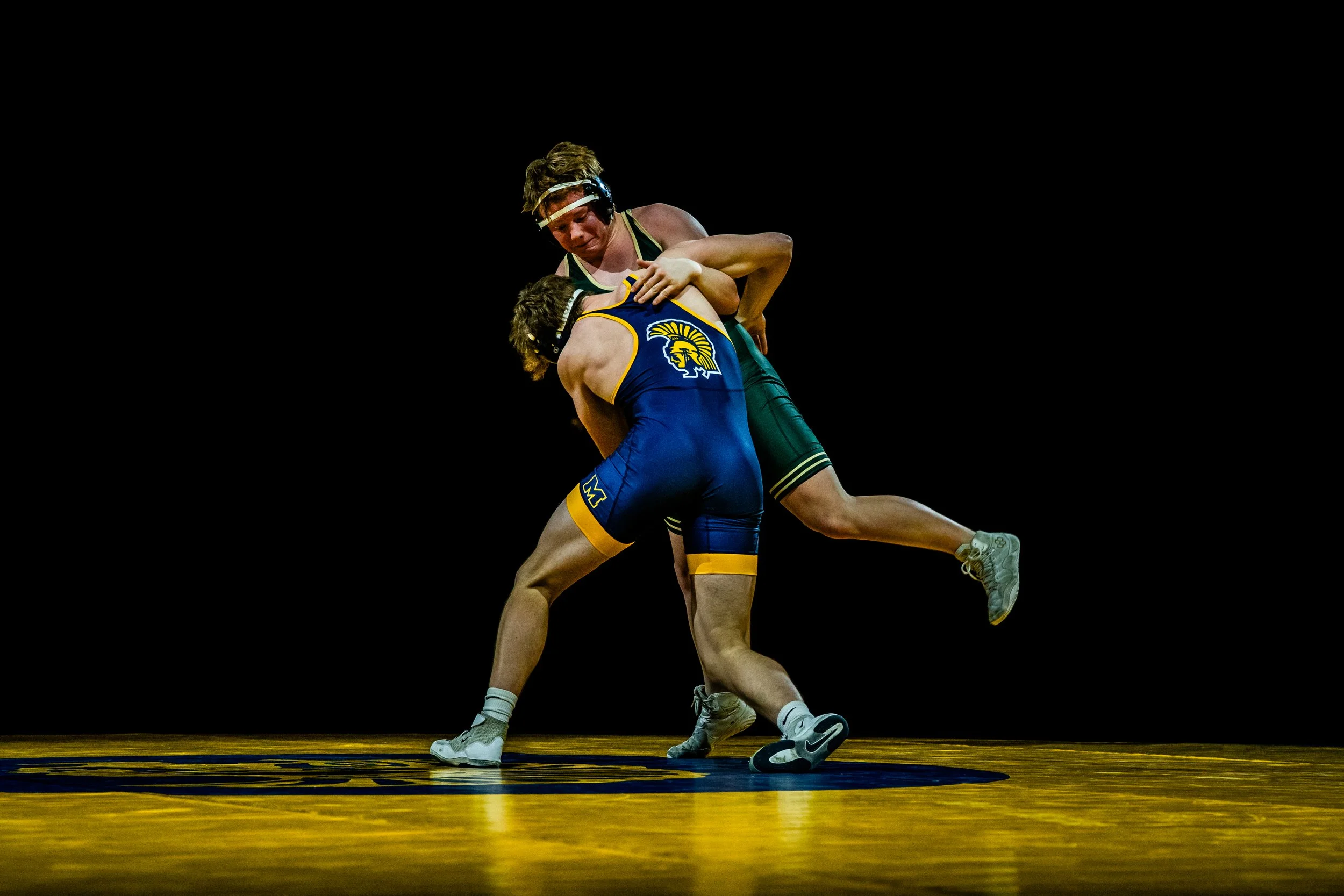 Two male wrestlers compete on a yellow wrestling mat; one wearing a blue singlet with yellow details, the other in a green singlet with black and white accents; both are wearing protective headgear.