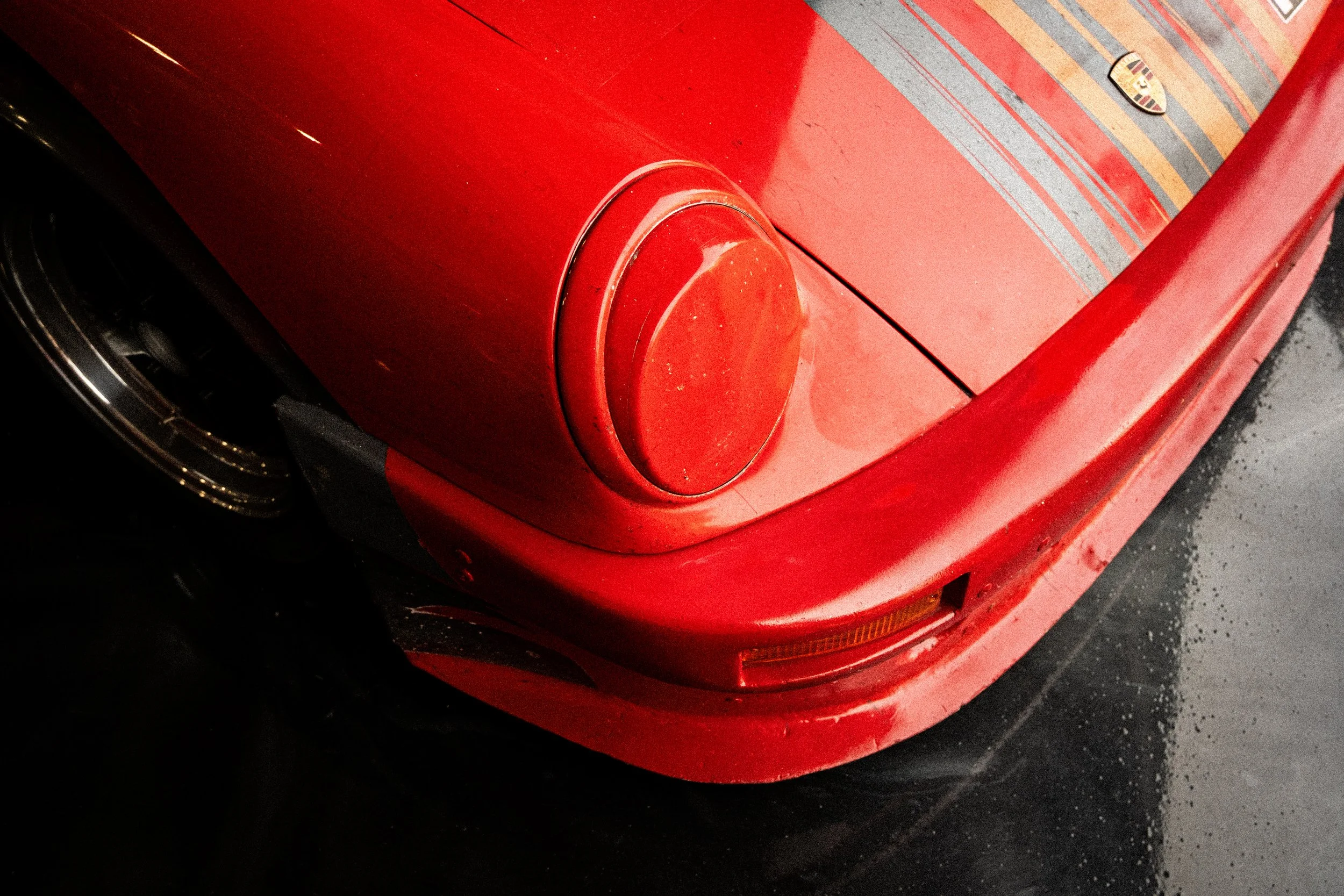 Close-up of the front part of a red sports car, highlighting the headlight and part of the hood, with a Porsche logo visible on the right side of the image.
