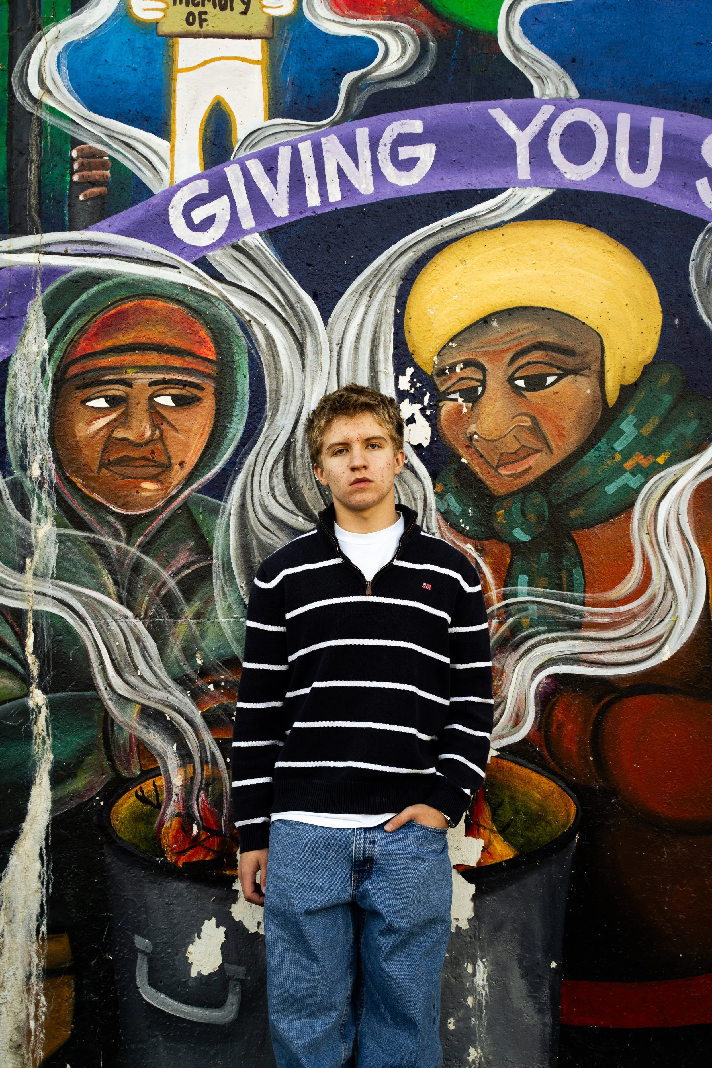 Young man standing in front of a colorful mural featuring two illustrated faces with a purple banner above reading 'Giving You'. The mural has vibrant colors and artistic details.