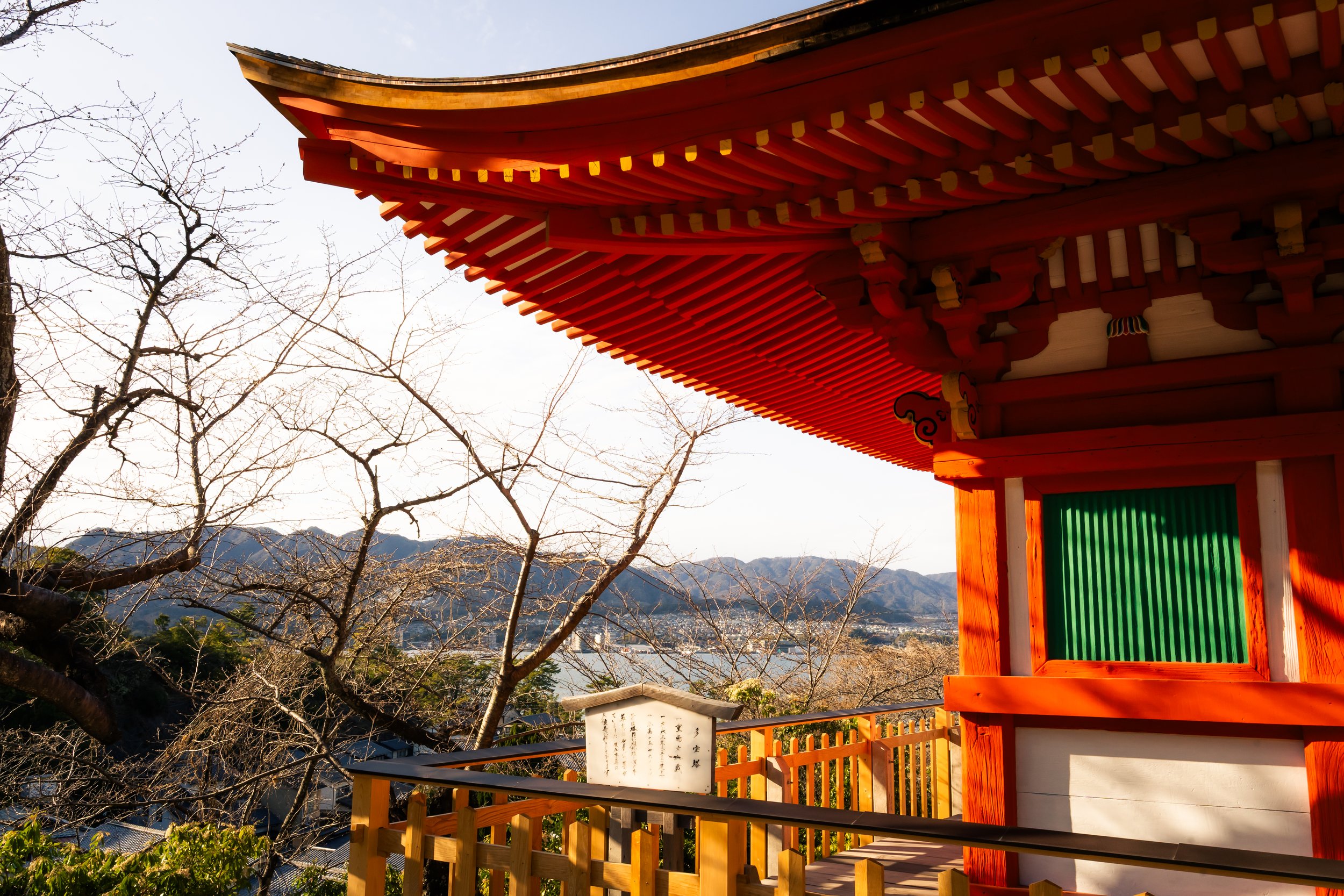 Traditional Japanese temple with a red wooden roof and green shoji screen, surrounded by leafless trees and mountains in the background.