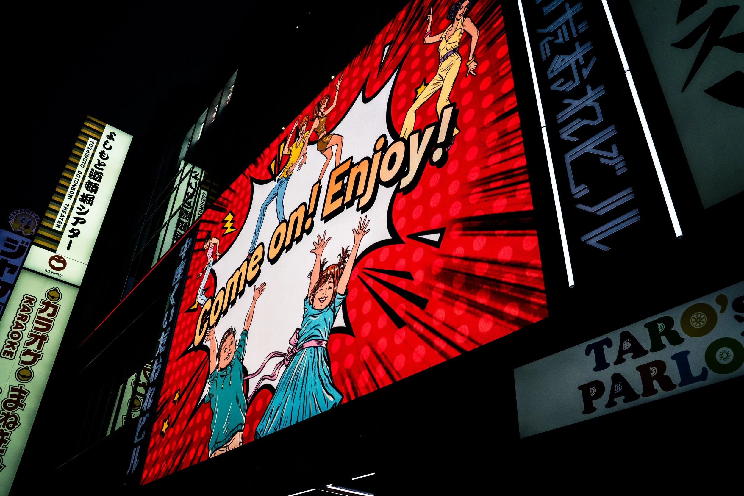 A large, colorful comic-style billboard with animated characters, primarily showing three children dressed in blue, enjoying themselves with energetic poses, and text in bold letters, set against a vibrant red background with dotted pattern.