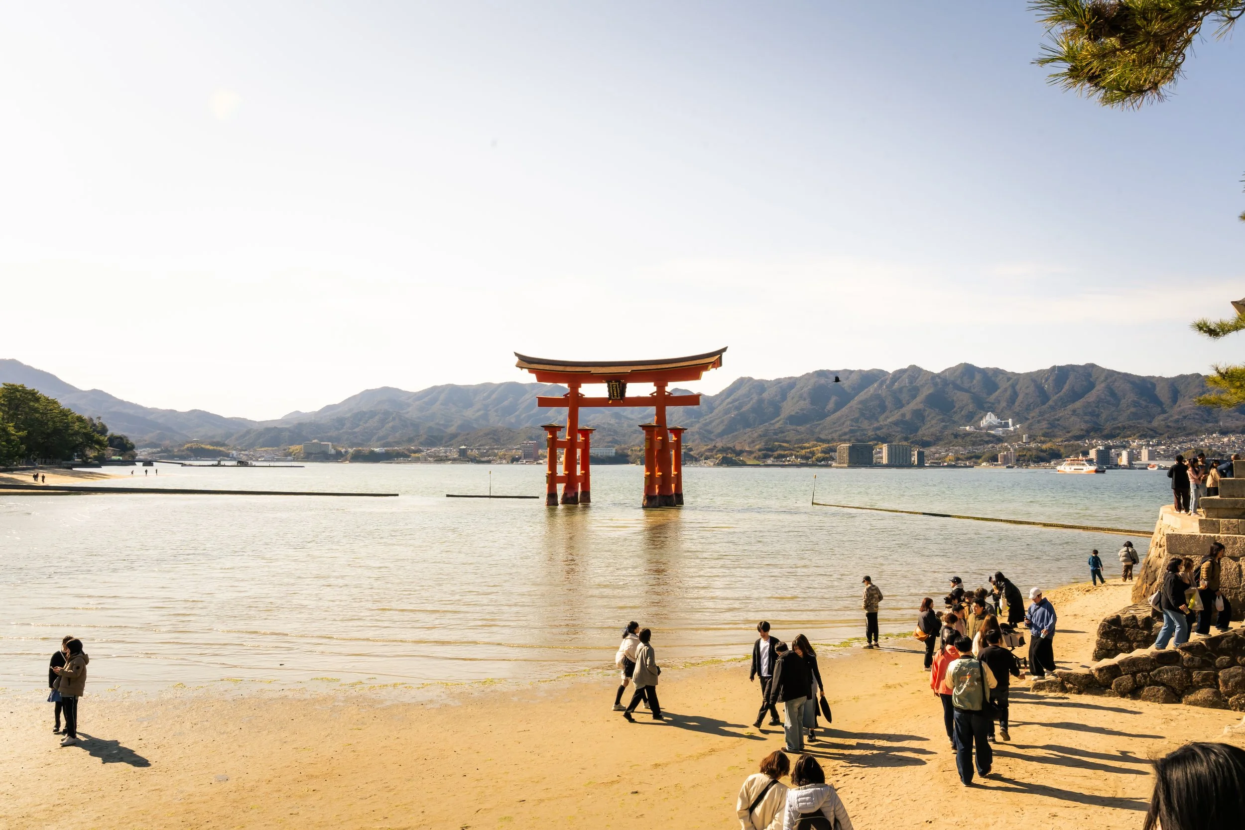 People walking along a beach with a large red torii gate in the water, mountains in the background, and a partly cloudy sky.