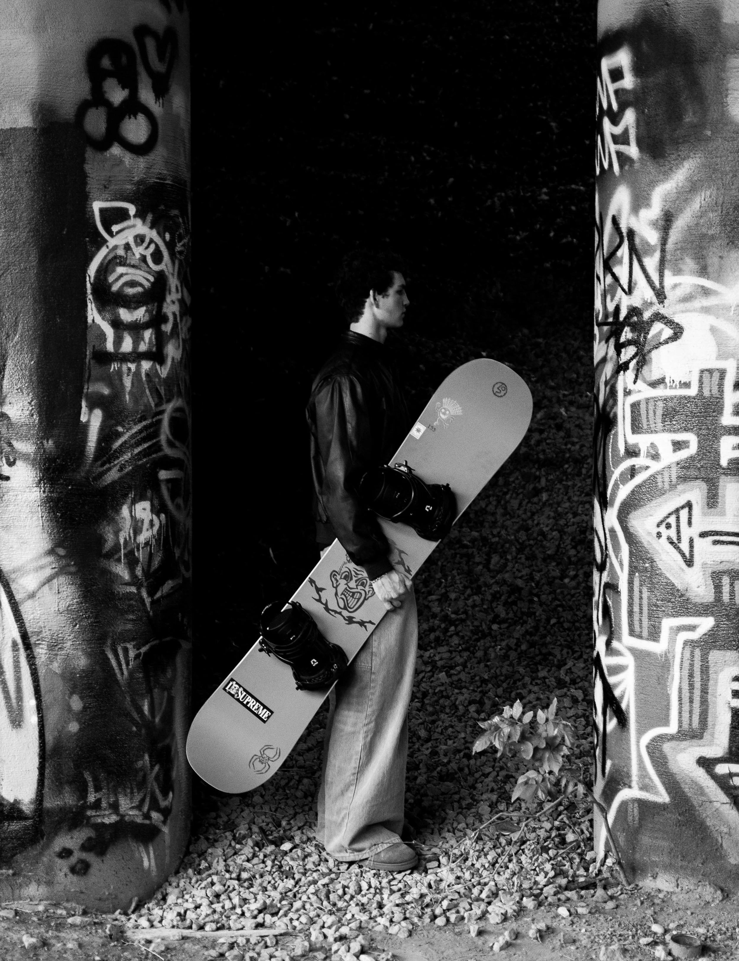 A person holding a skateboard stands between two graffiti-covered pillars in an outdoor setting with gravel ground.