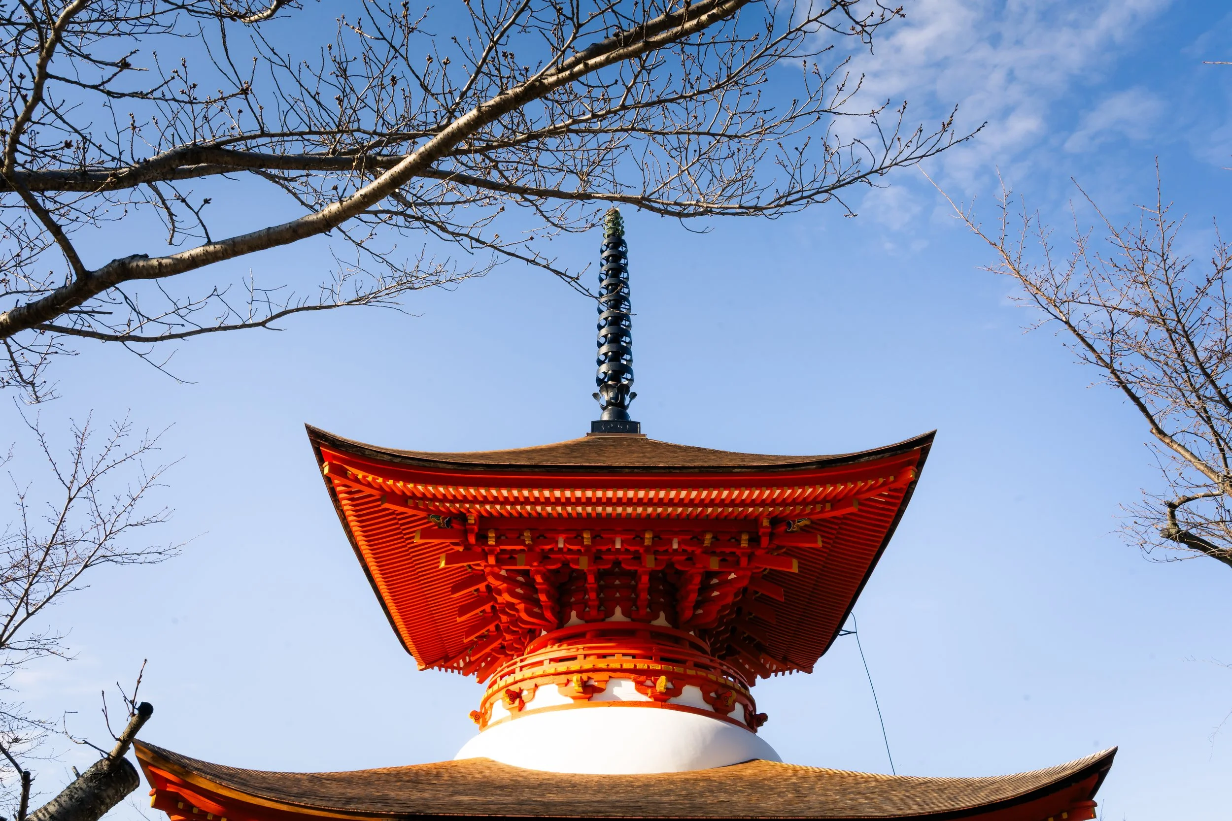 Traditional Japanese pagoda with a red and orange roof, fine black finial, and leafless tree branches in the foreground against a clear blue sky.