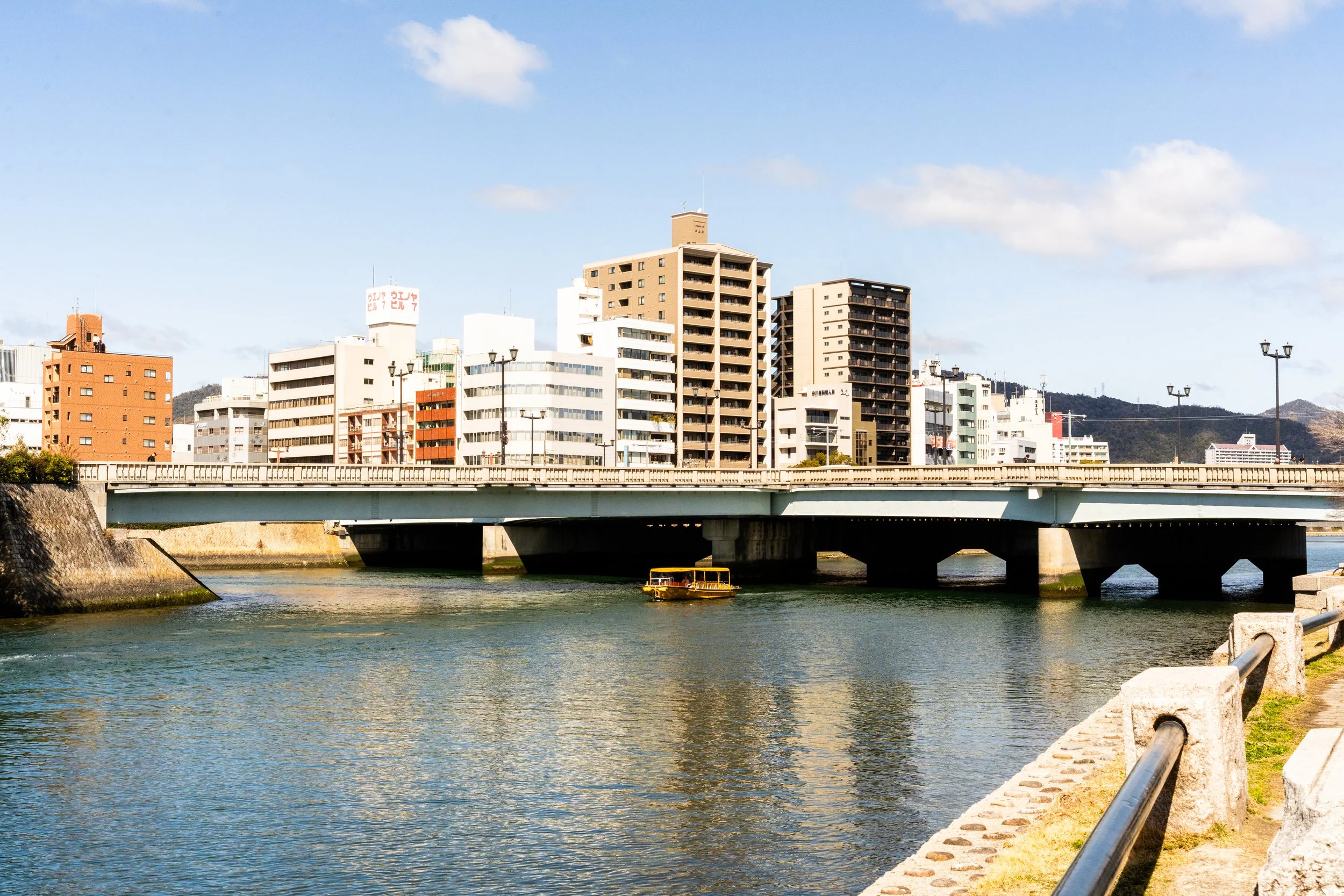 Cityscape of high-rise buildings along a river with a bridge, a small yellow boat, and a walkway with a metal railing.