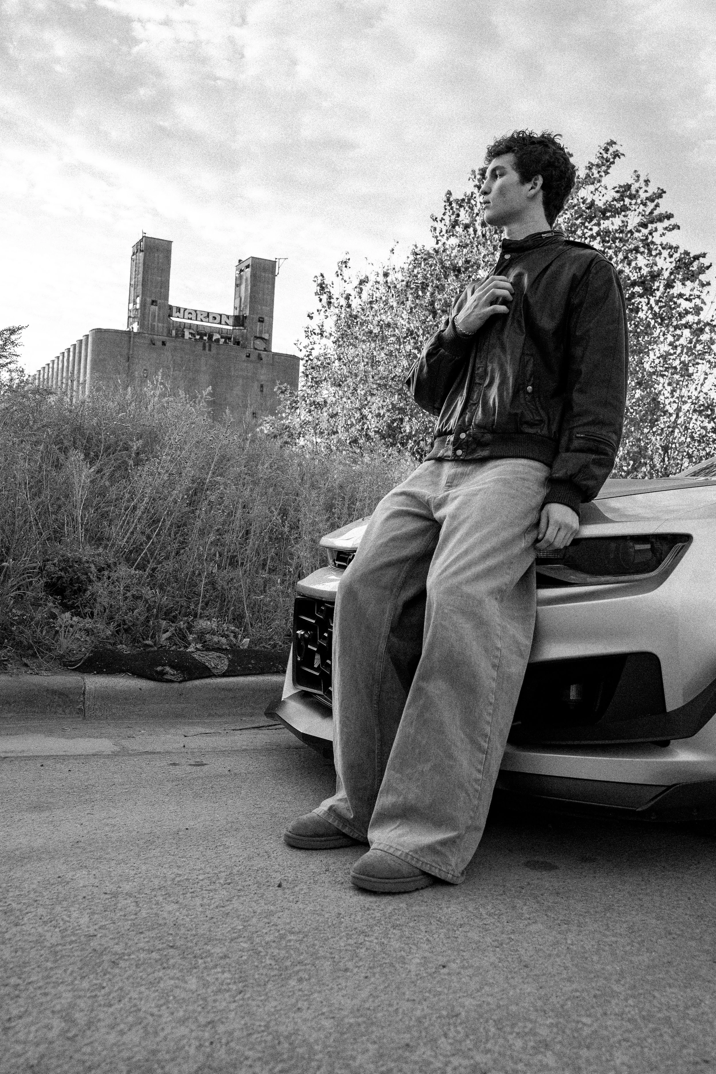 A young man with curly hair standing beside a car, leaning against it with one hand on his chest, outdoors with trees and an industrial building in the background.