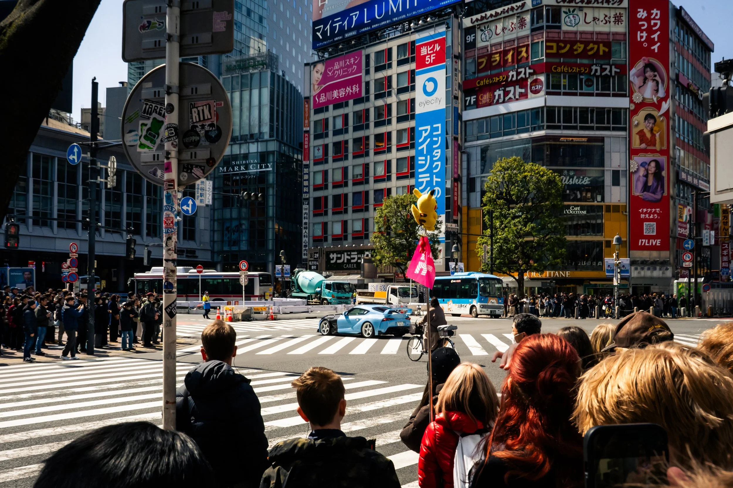 Crowd of people waiting to cross the street at a busy intersection in an urban city, with tall buildings and colorful advertisements and signs in the background.