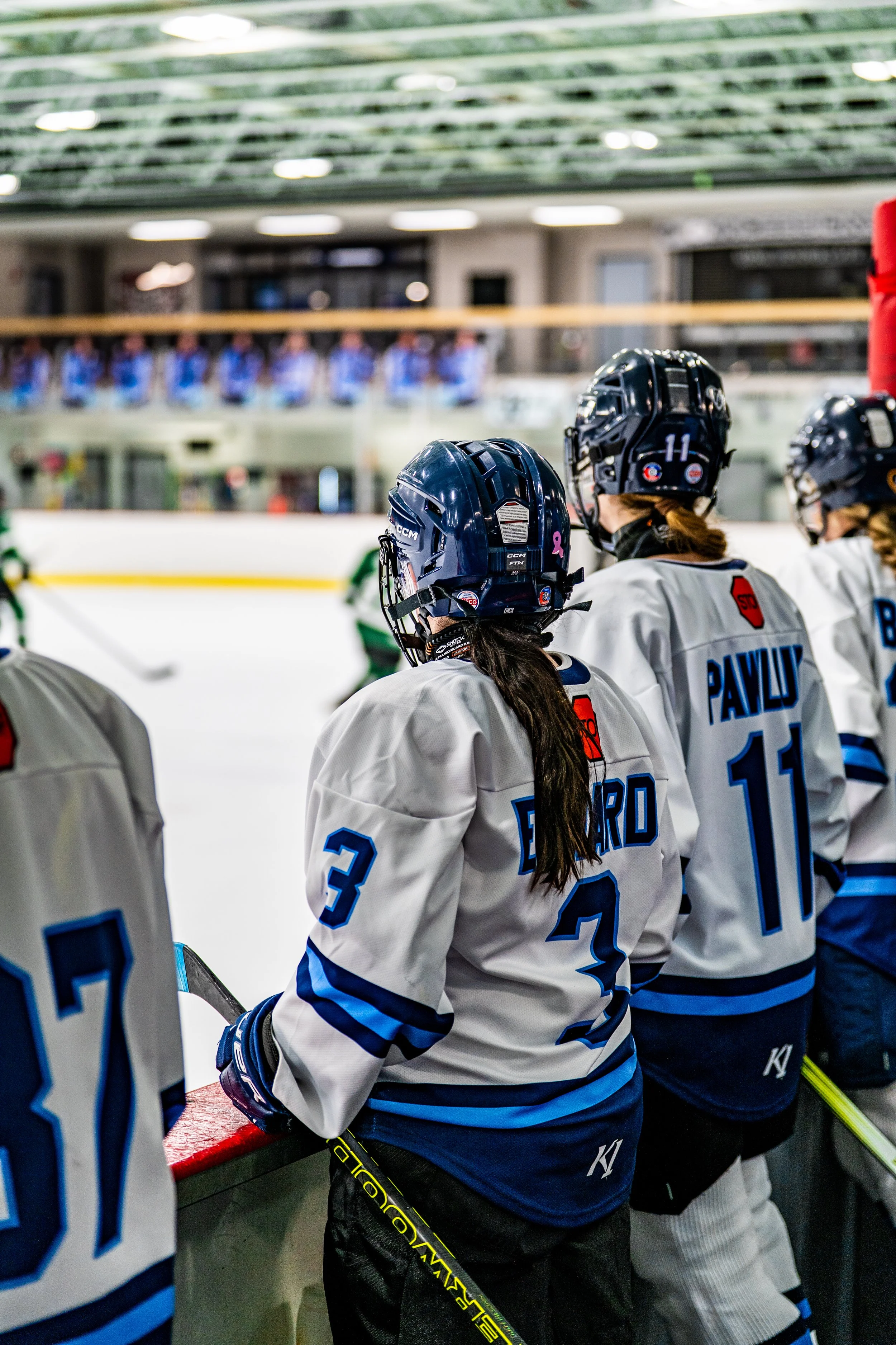 Hockey players in white jerseys with blue accents standing on the ice rink, wearing black helmets, during a game or practice.