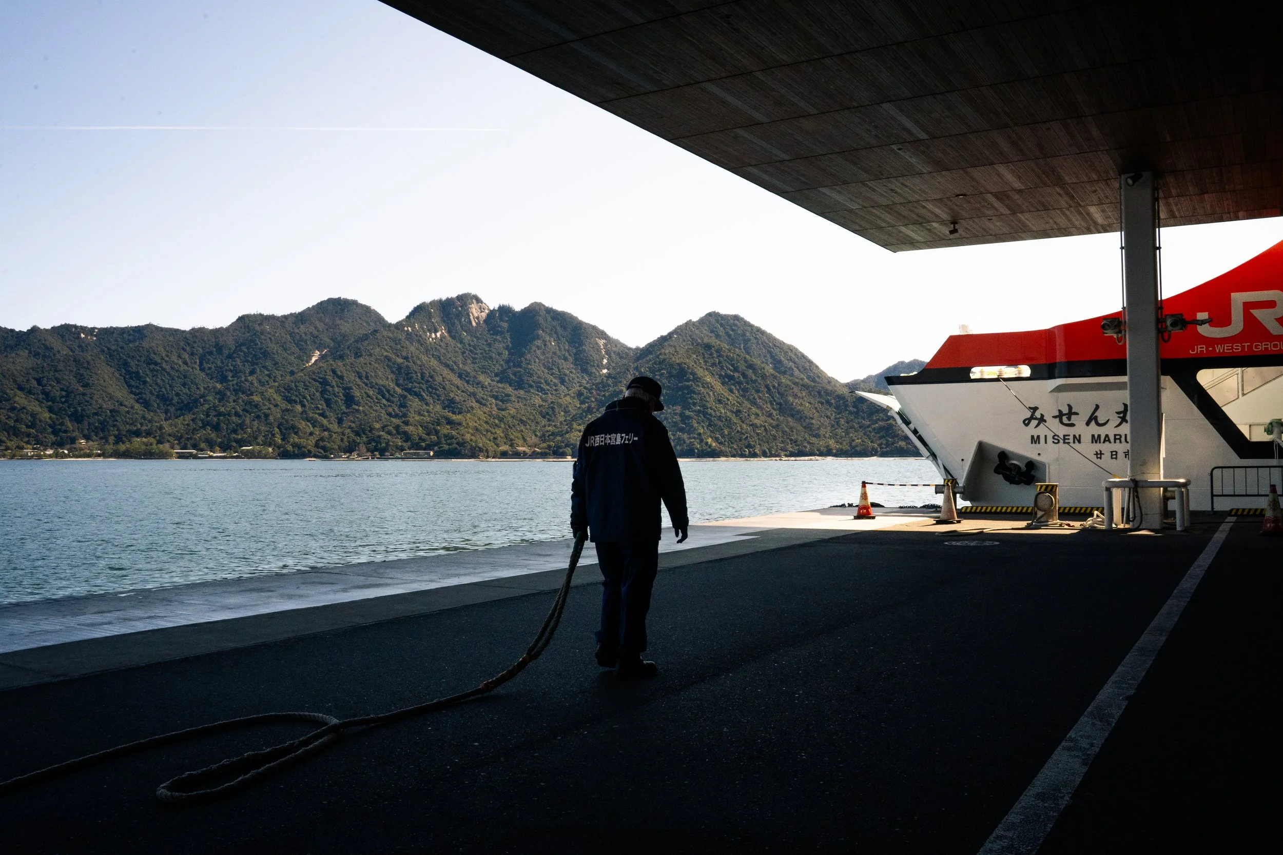 A person standing on a dock with a large boat named Misen Maru docked beside them. The person is holding a thick rope, and there are scenic mountains in the background across a body of water. The dock is shaded under a building's overhang, and it app