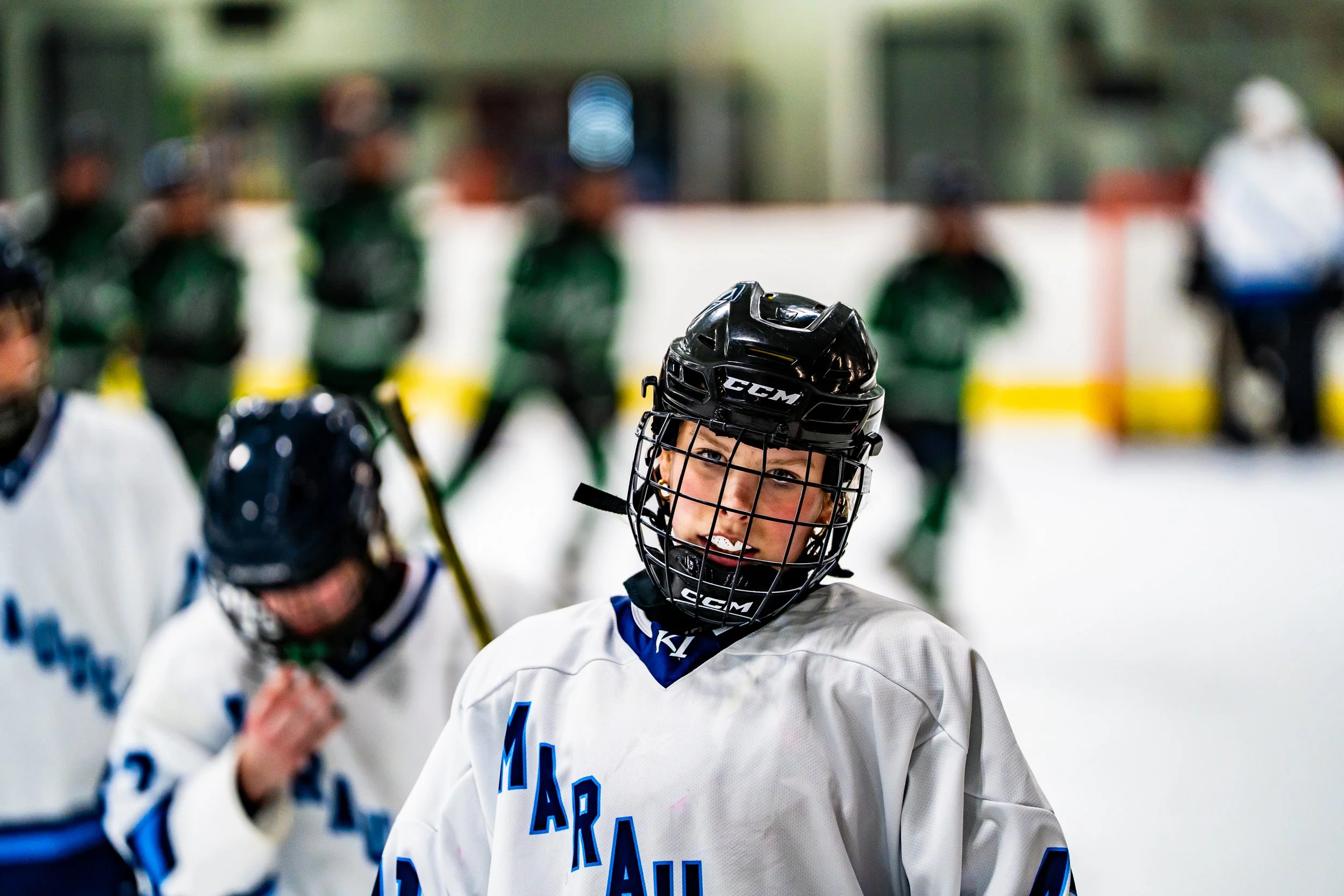 A young hockey player in a black helmet and white jersey with blue lettering, smiling at the camera inside an ice rink, with teammates and coaches in the background.