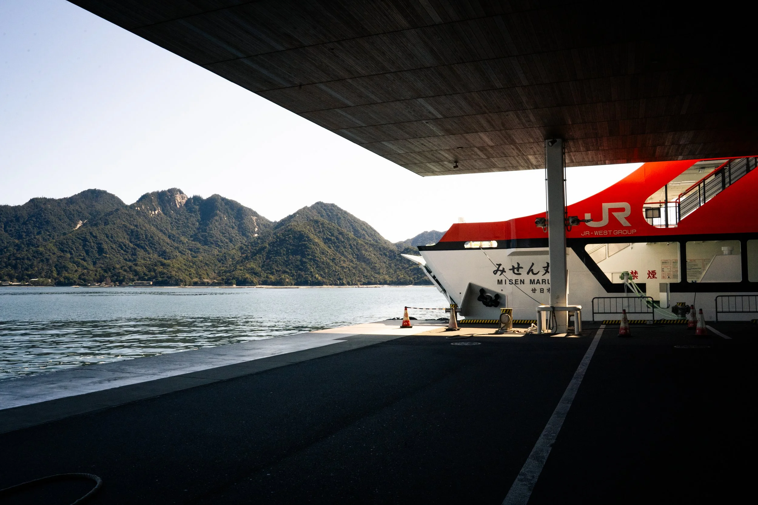 Ferry docked at a waterfront with mountains in the background, partly shaded by a large overhang.