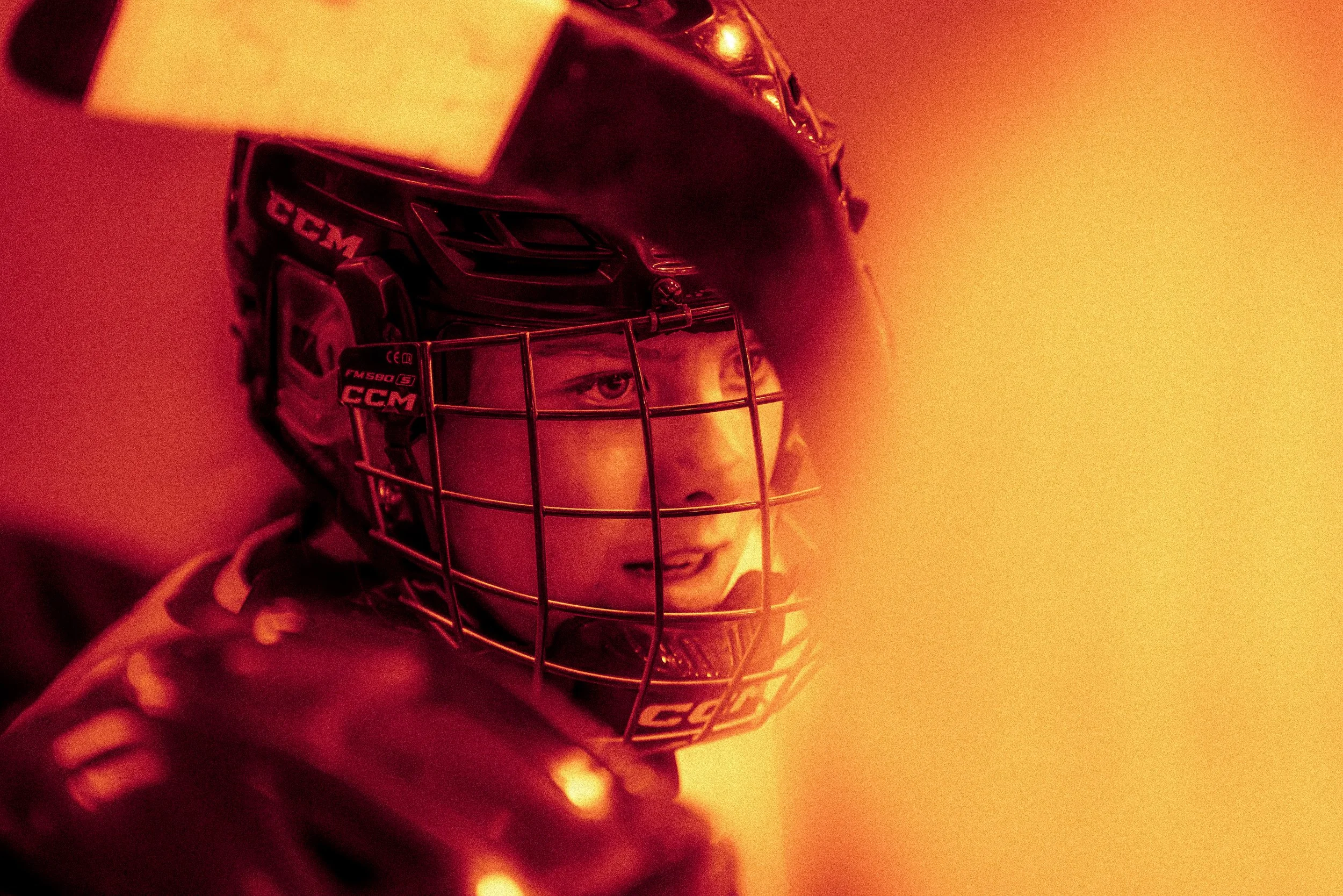 Close-up of a young hockey player wearing a black helmet with a face cage, with intense focus and determination in their eyes.