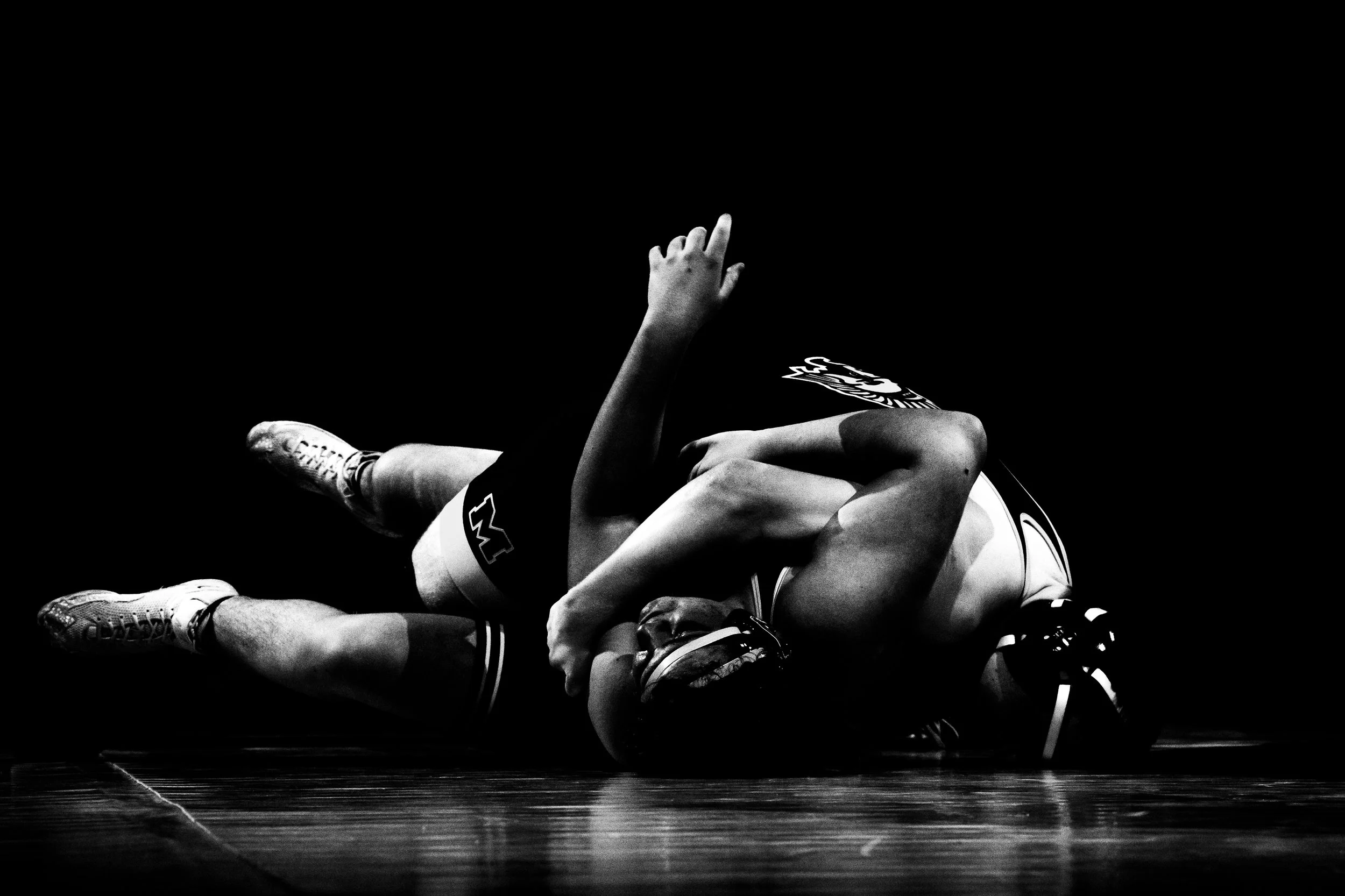 Two wrestlers on the mat, one on top of the other, engaged in a match. The image is in black and white.