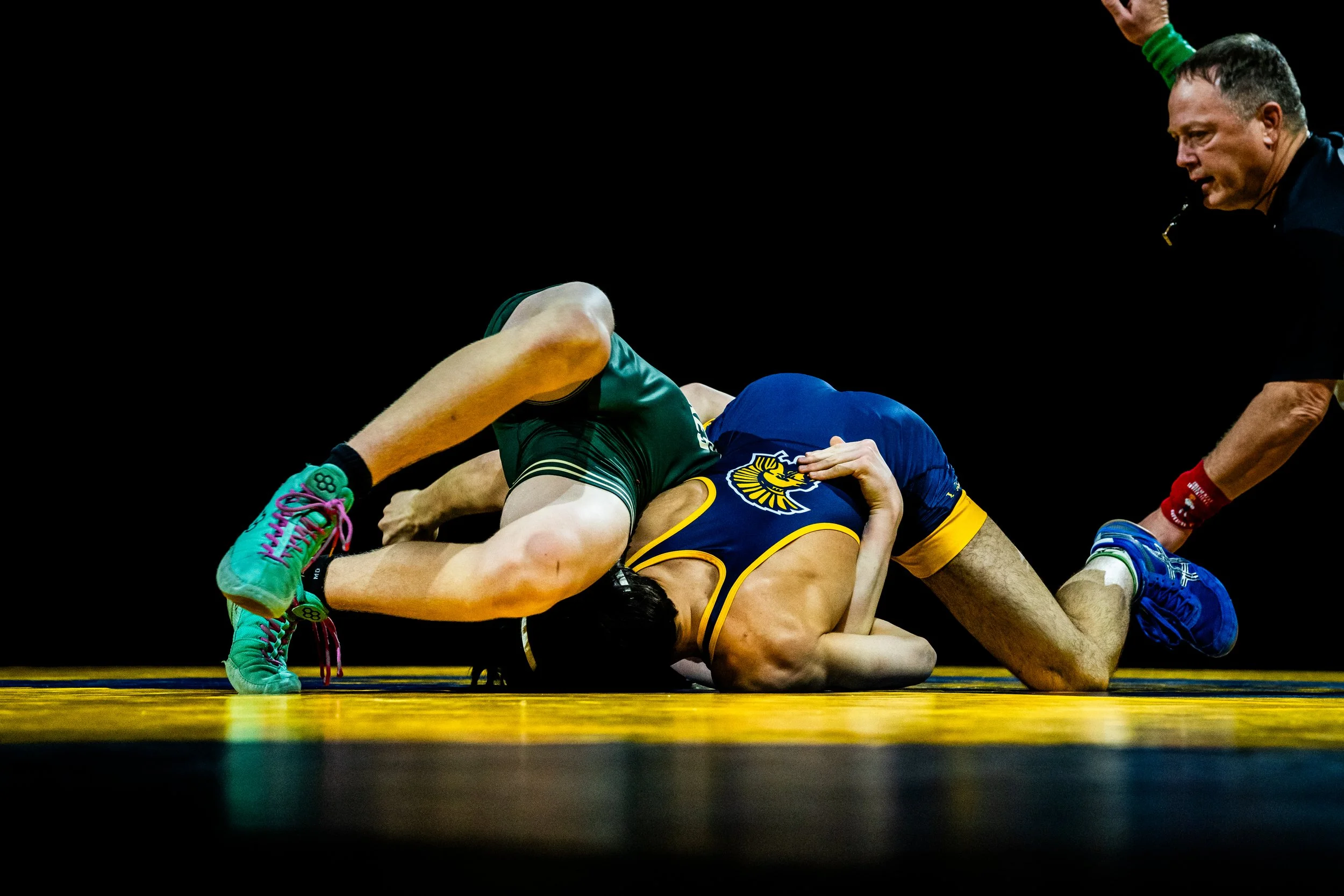 Wrestlers on the mat during a match, one in a green outfit and the other in a yellow and blue outfit, with a referee nearby.