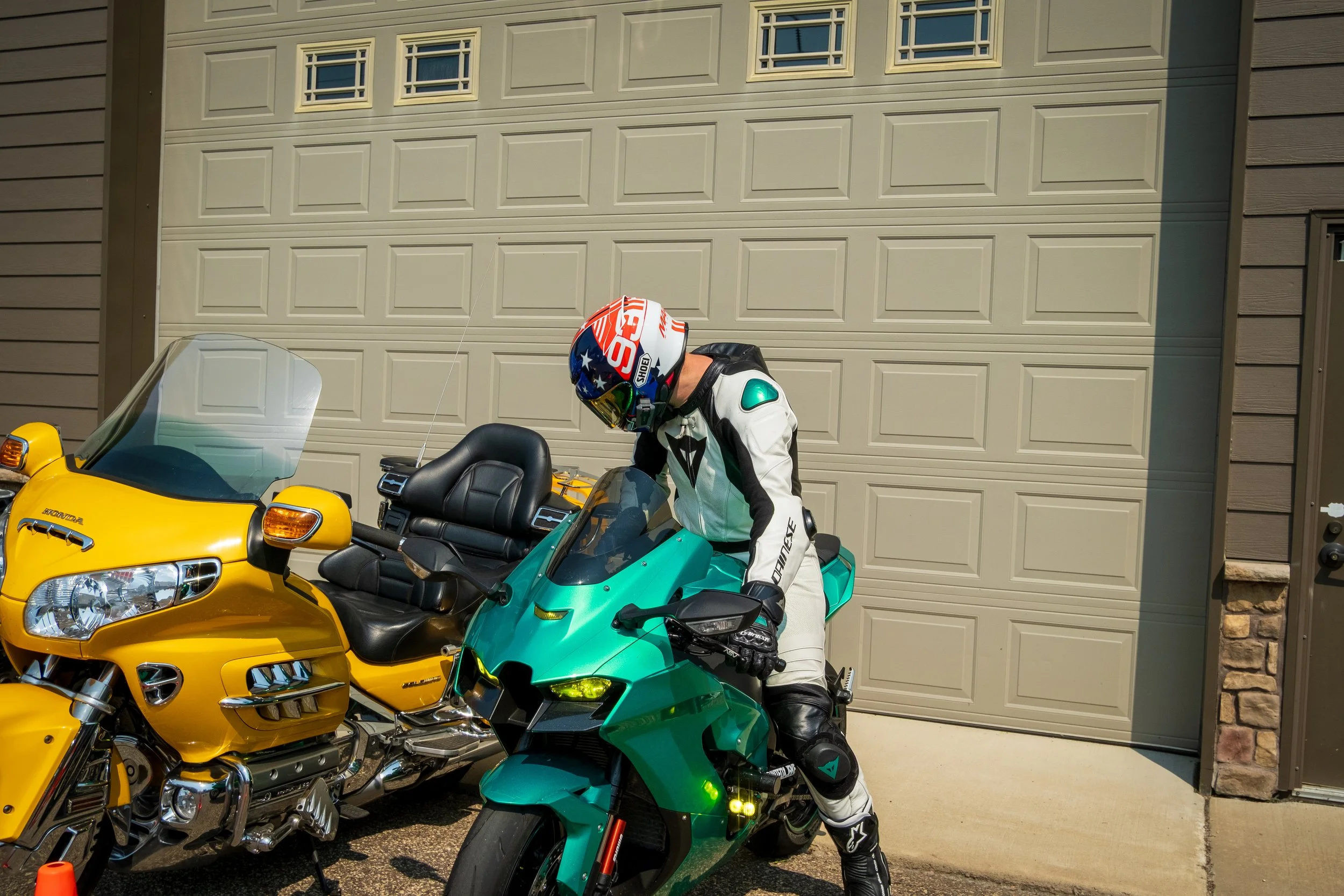 A person dressed in motorcycle gear, including a colorful helmet, white riding suit, and black boots, sitting on a green motorcycle parked in front of a beige garage door. Two yellow motorcycles are parked beside the green motorcycle. The scene is ou