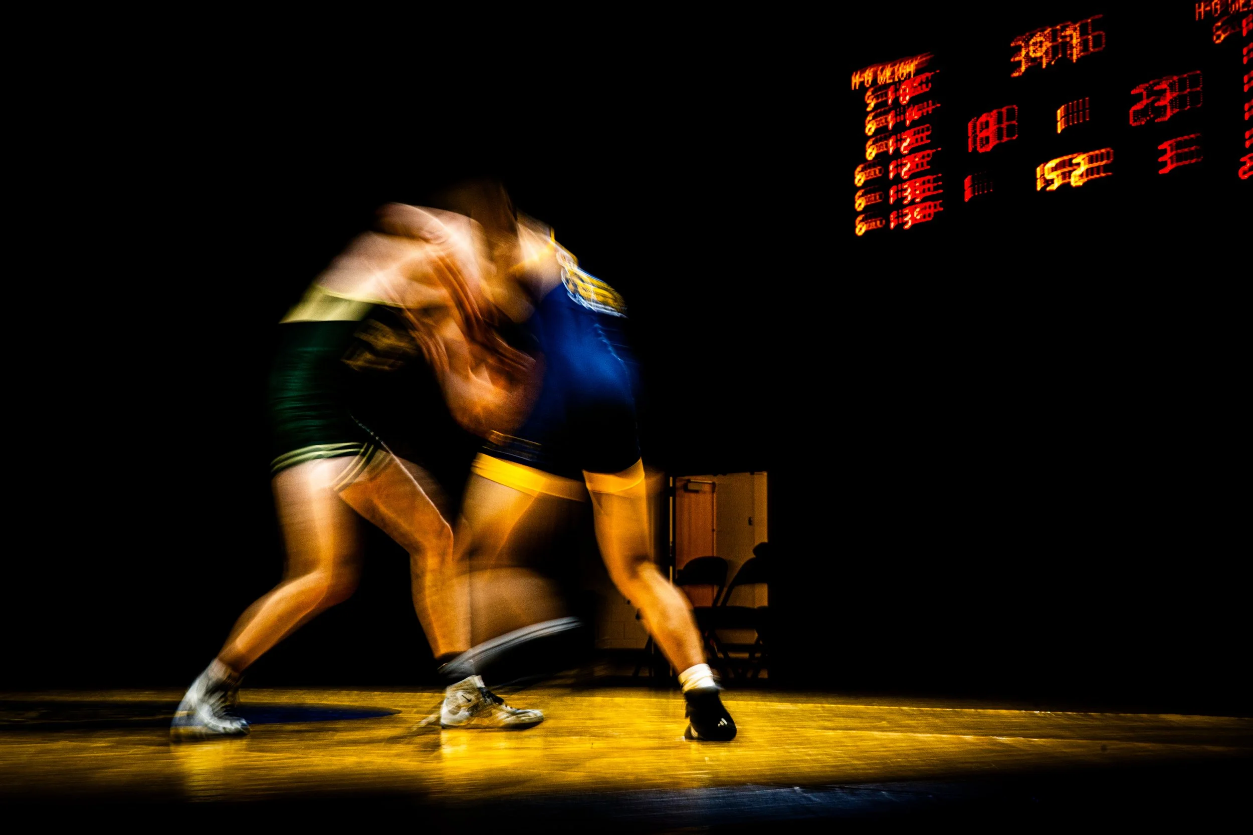 Two wrestlers in black and yellow uniforms wrestling on a wooden mat in a dimly lit gym, with an electronic scoreboard showing red numbers in the background.