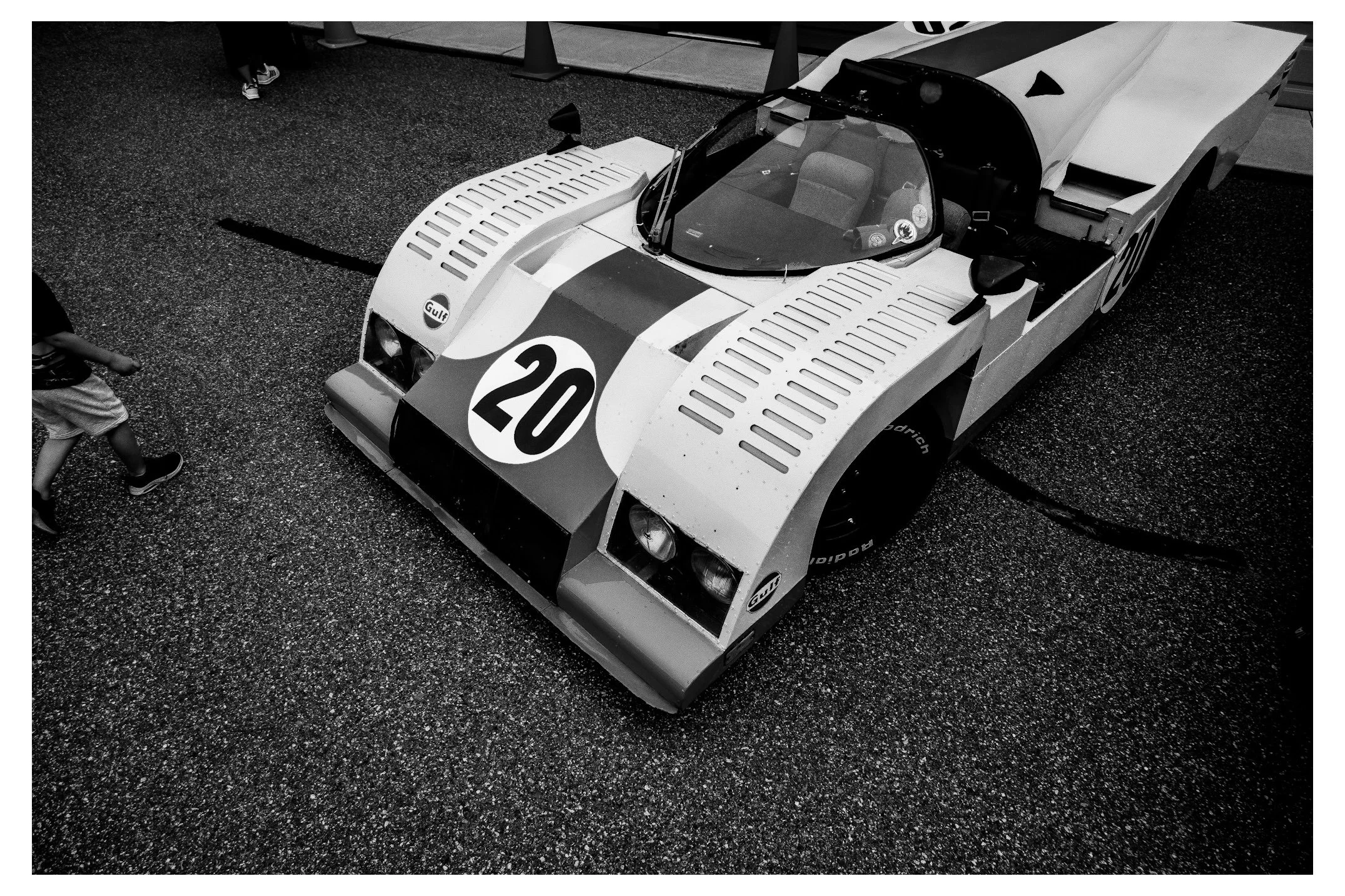 A vintage race car with the number 20 on the front and Gulf logo, parked on a textured surface within a barricaded area at a racing event, with a person in shorts walking nearby.