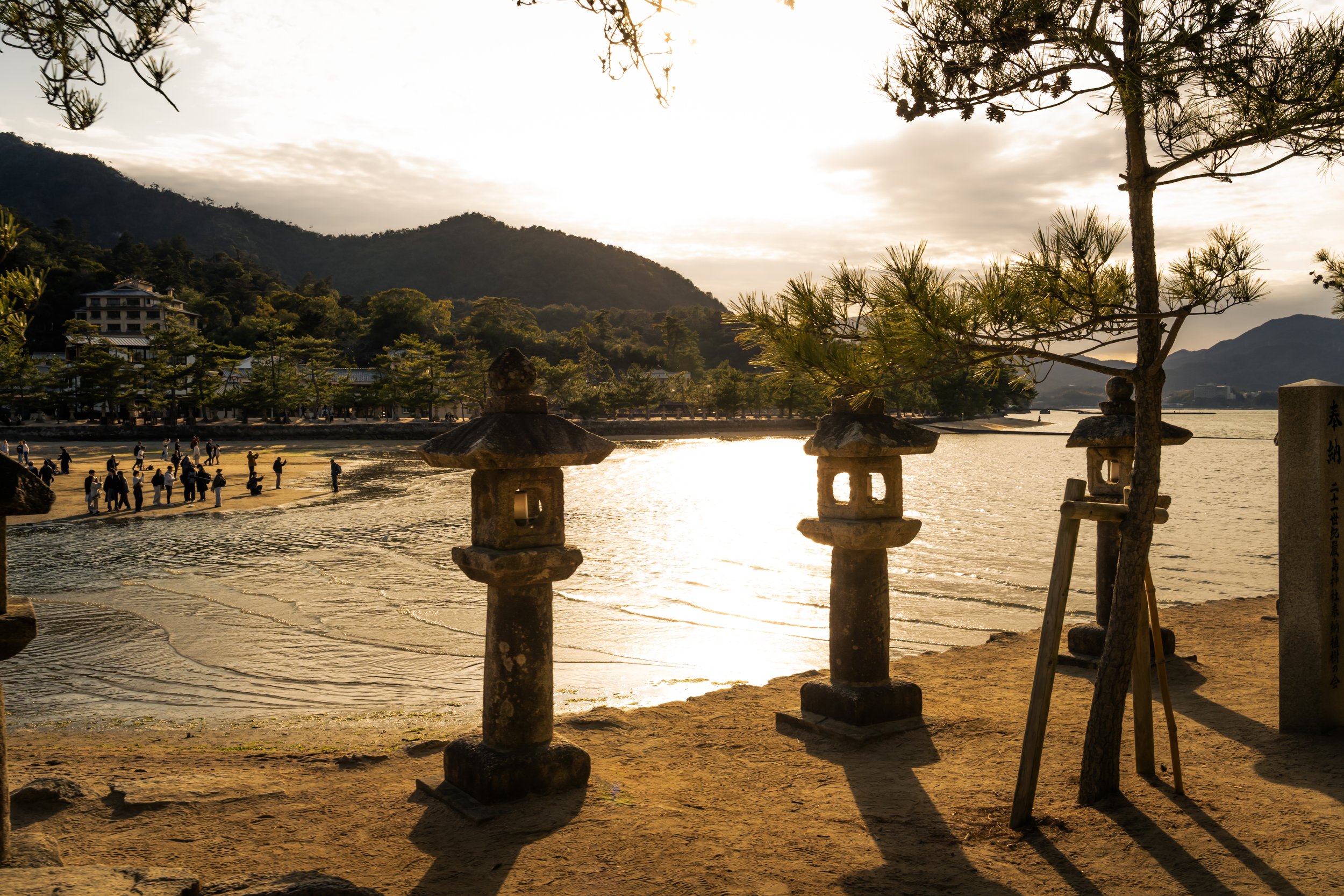 A lakeside scene at sunset with trees, mountains, and a group of people walking along the shore. Traditional stone lanterns and a pine tree are in the foreground.