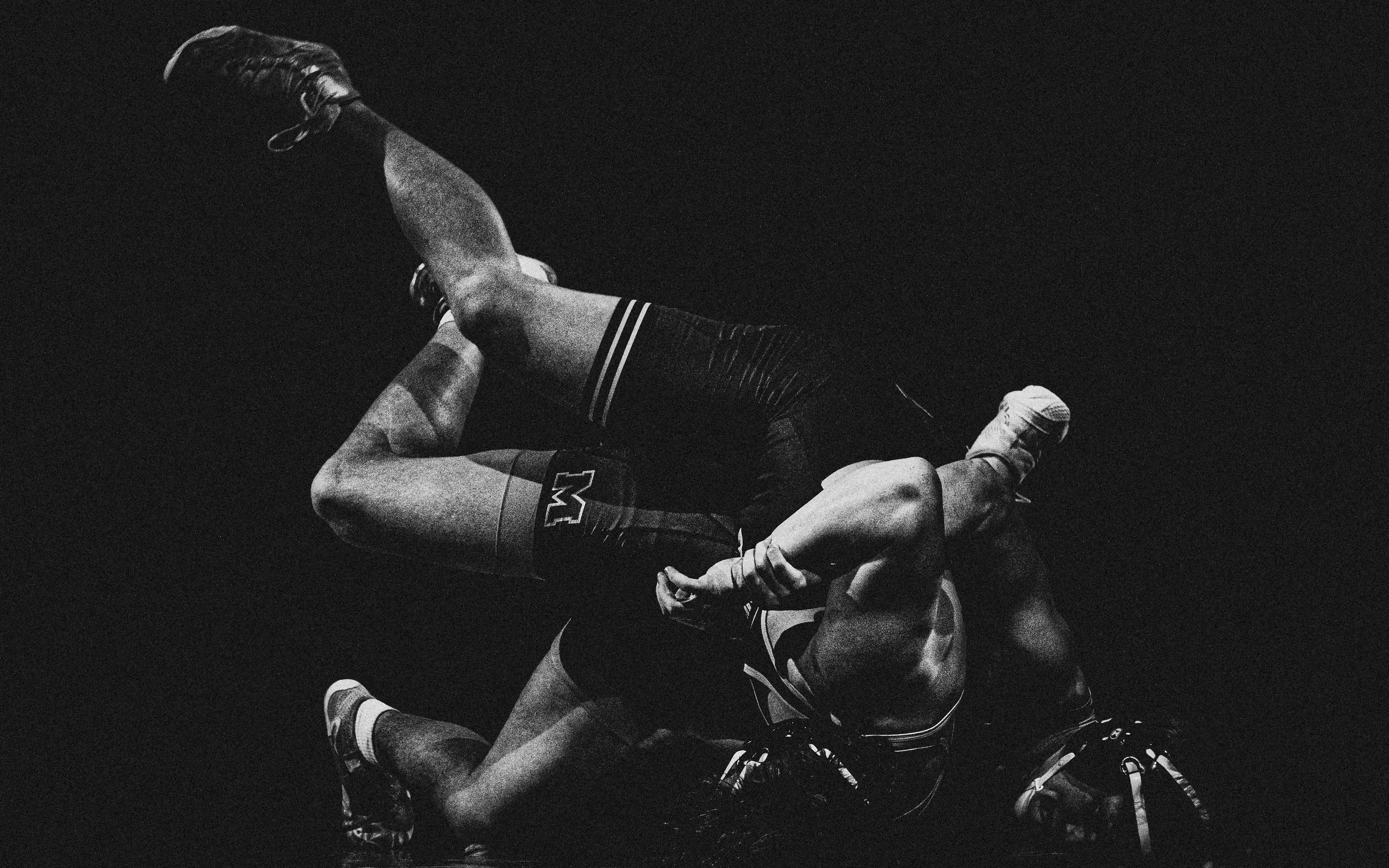 Two male wrestlers engaged in a wrestling match, with one wrestler on top and the other on the bottom, on a dark background.